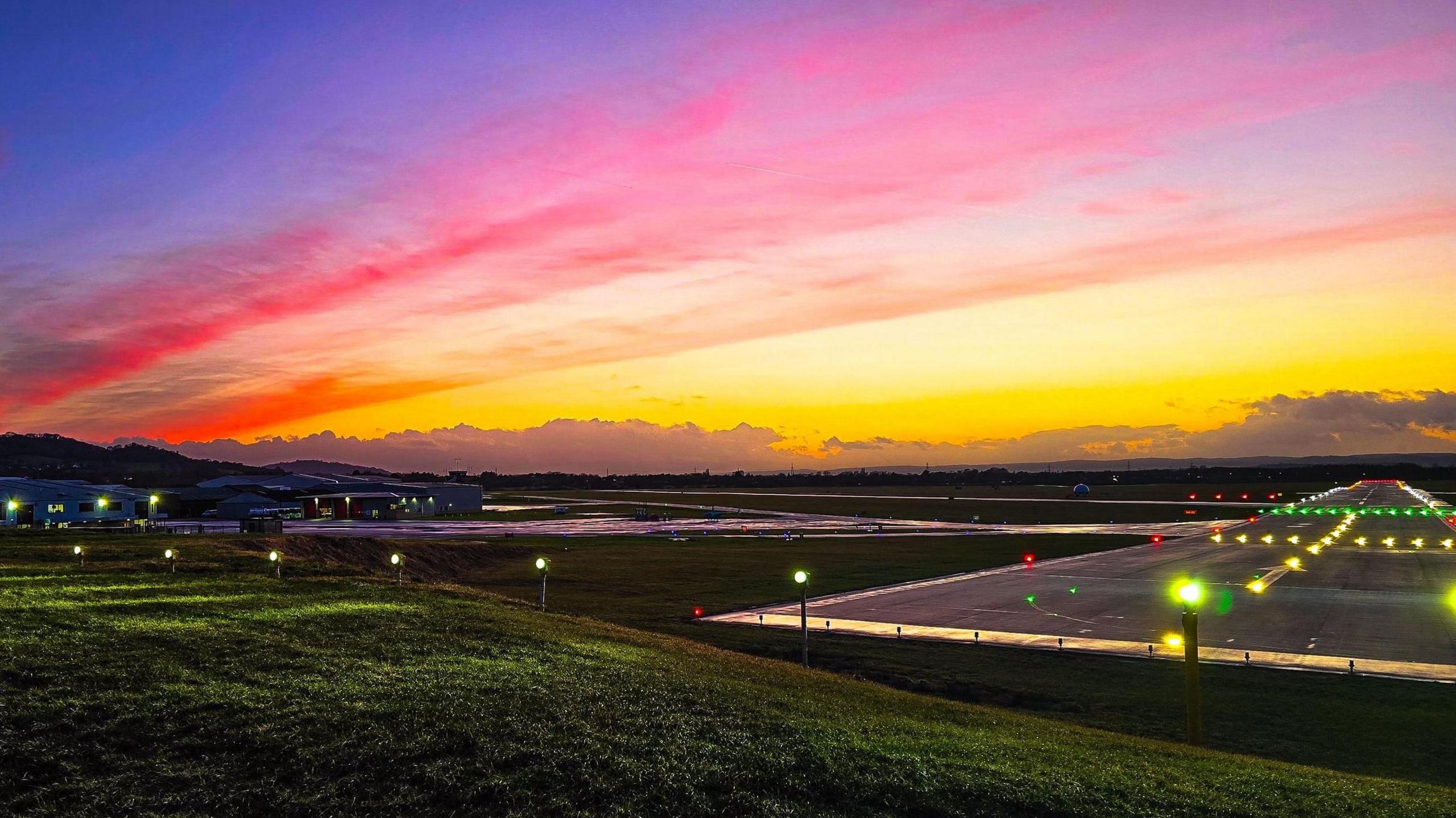 A photo of Gloucestershire Airport with the sunset in the background of the image. The colours range from orange, to pinks, to yellows. 