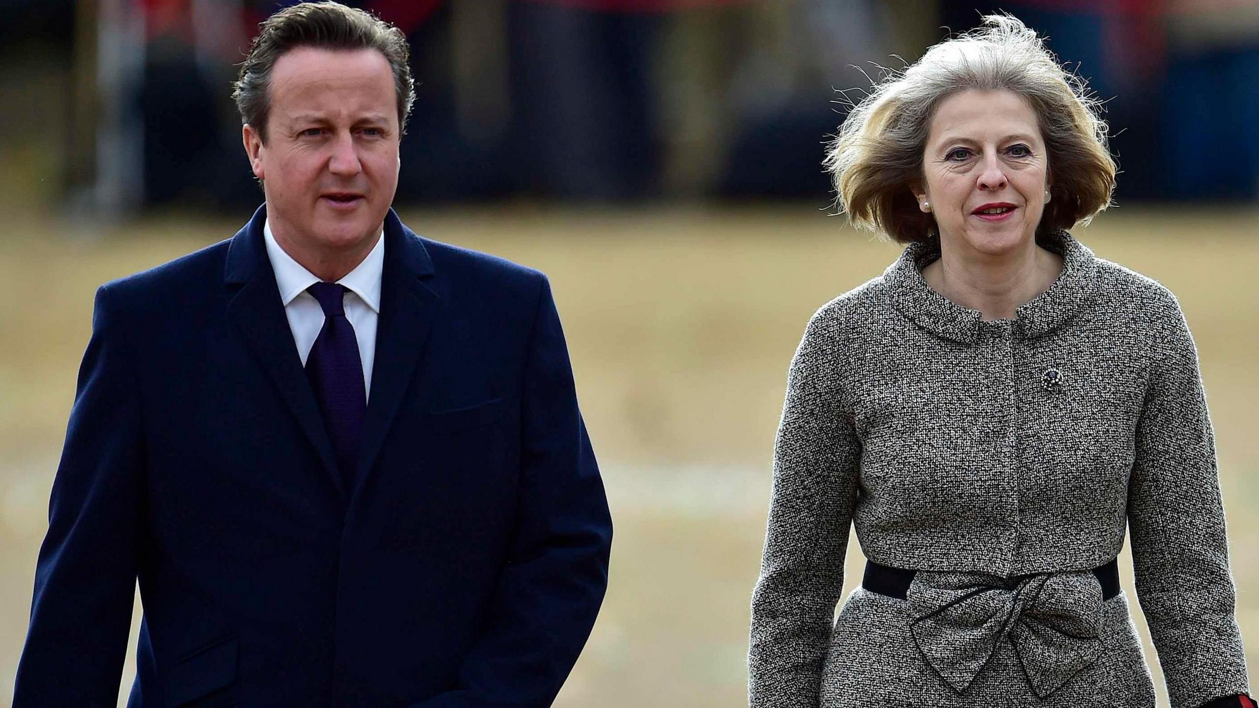 Lord David Cameron in a blue overcoat, white shirt and blue tie, walks alongside Baroness Theresa May, who wears a grey and white patterned jacket with a large bow belt