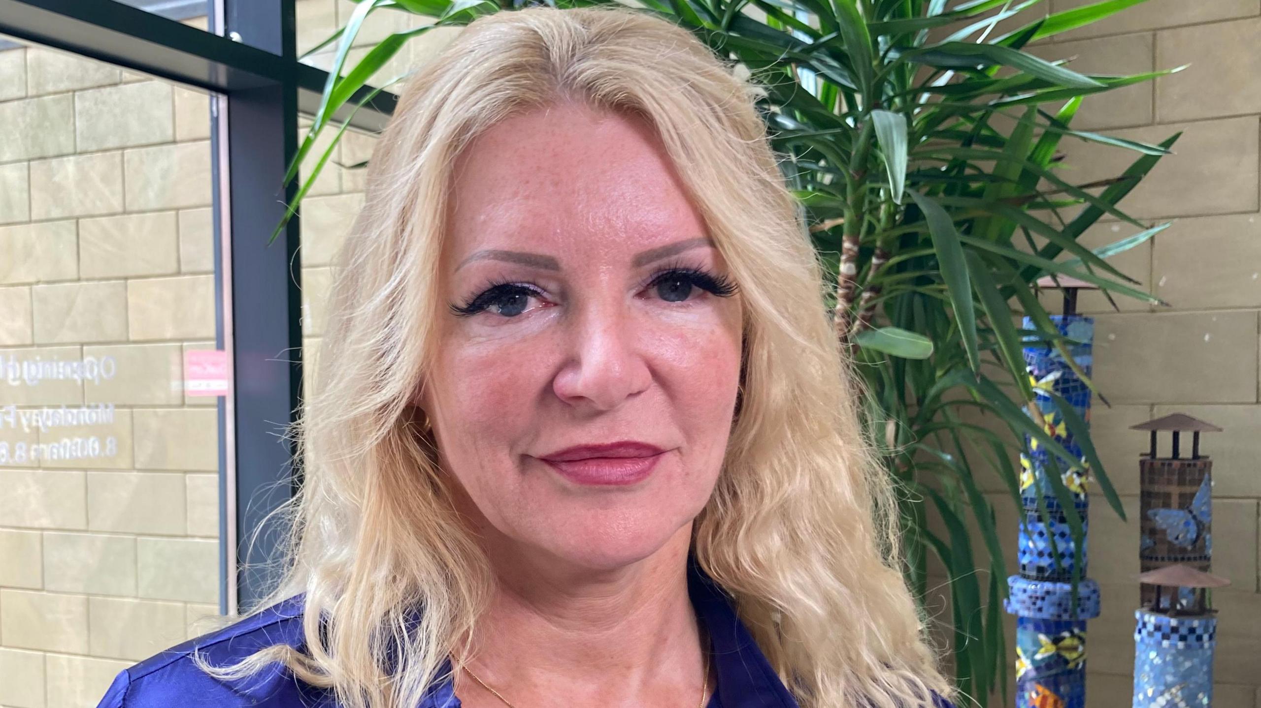 Mechelle Lewis has long blonde hair and is wearing a blue top and a gold chain round her neck. Behind her is a glass window and a large green plant.