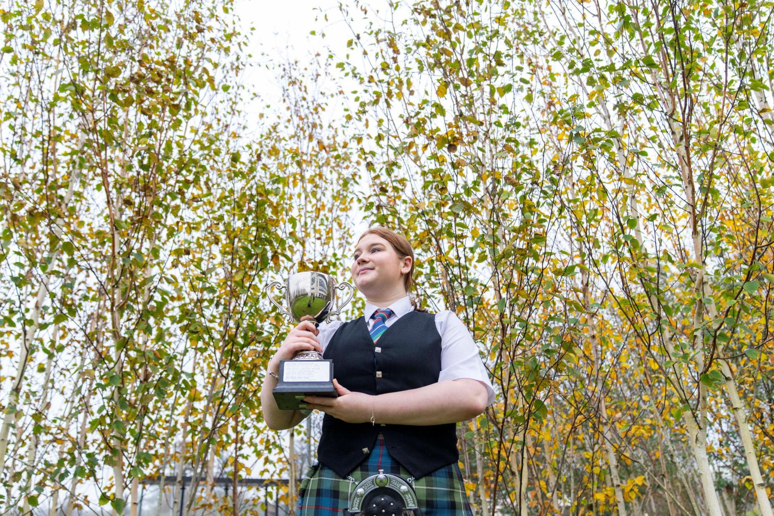 Lily Robertson is pictured outside among silver birches with their leaves turning an autumnal yellow and gold. Lily is wearing a green and blue tartan kilt and a black waistcoat over a white shirt. Her brown hair is tied back and she is smiling as she holds her trophy.