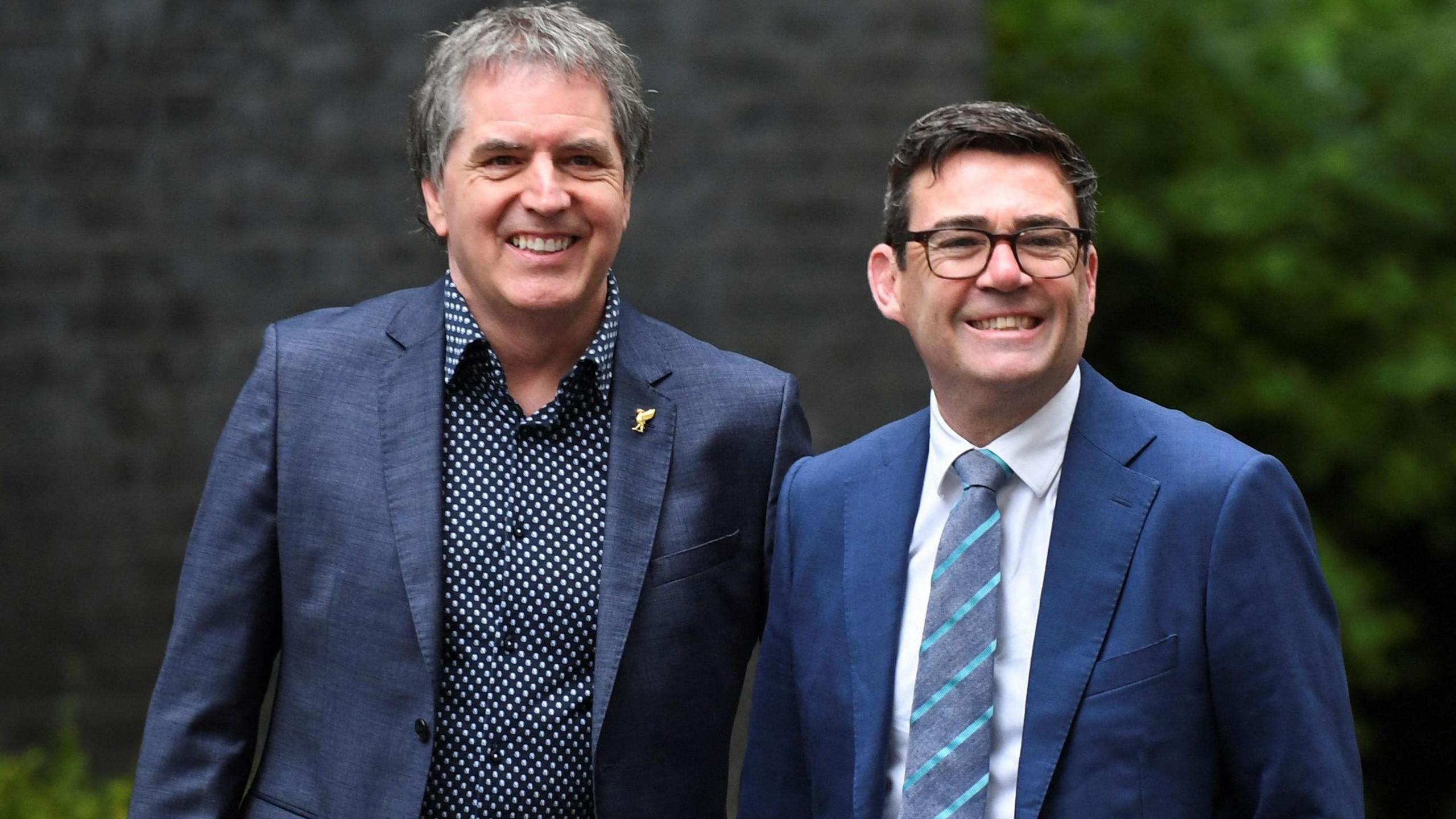 Steve Rotheram has greying hair is dressed in bluey grey suit and dark blue shirt with white polka dots is on the left with Andy Burnham in lighter blue suit, white shirt he has dark brown hair, dark brown spectacles and a blue striped tie