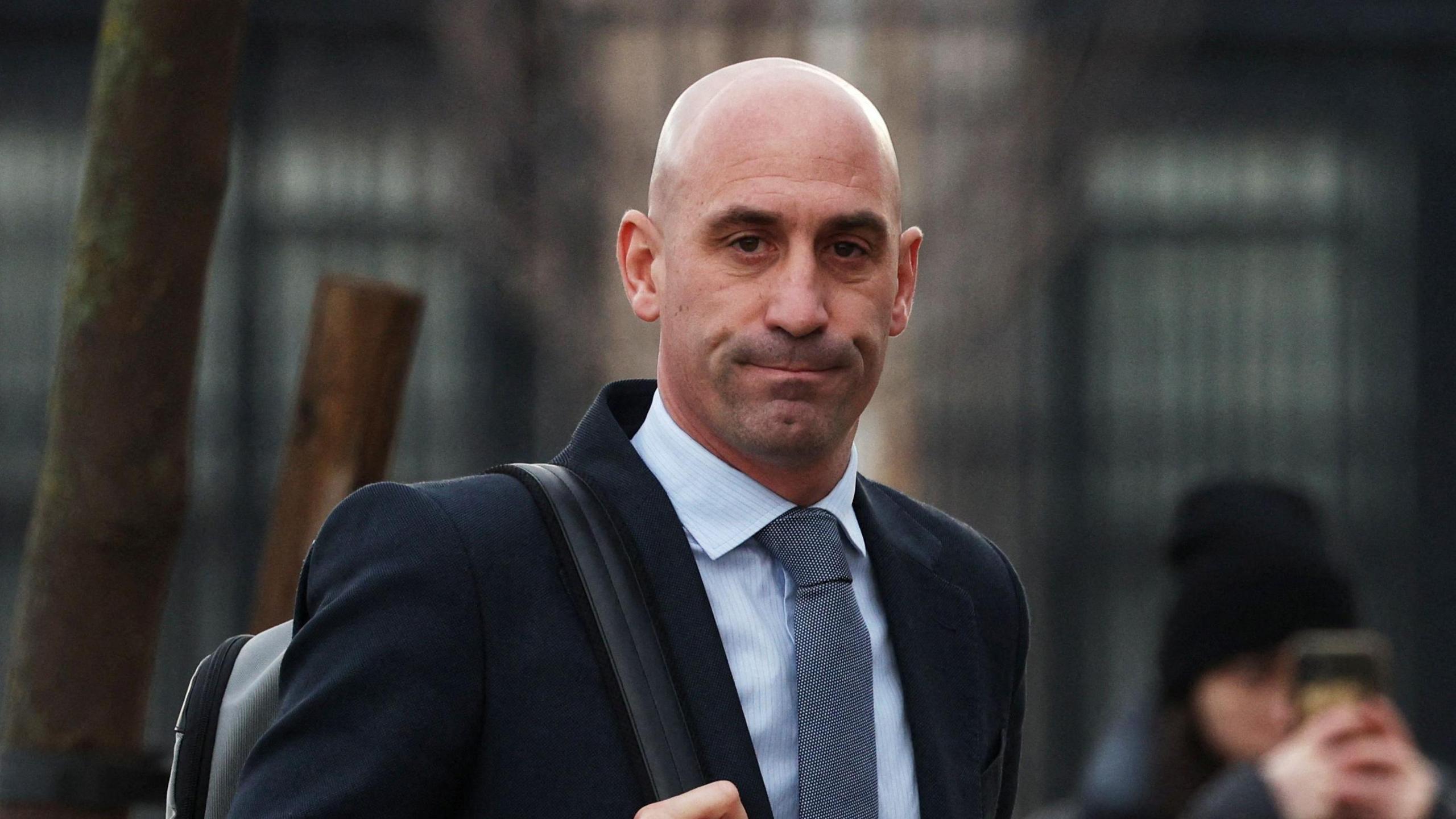 Luis Rubiales hold the strap of the backpack his is wearing over his right shoulder. He is wearing a suit and tie and is outside, on his way to the court in February 2025. In the background is a blurry woman wearing a beanie hat, holding her mobile phone up to her face to take a picture.