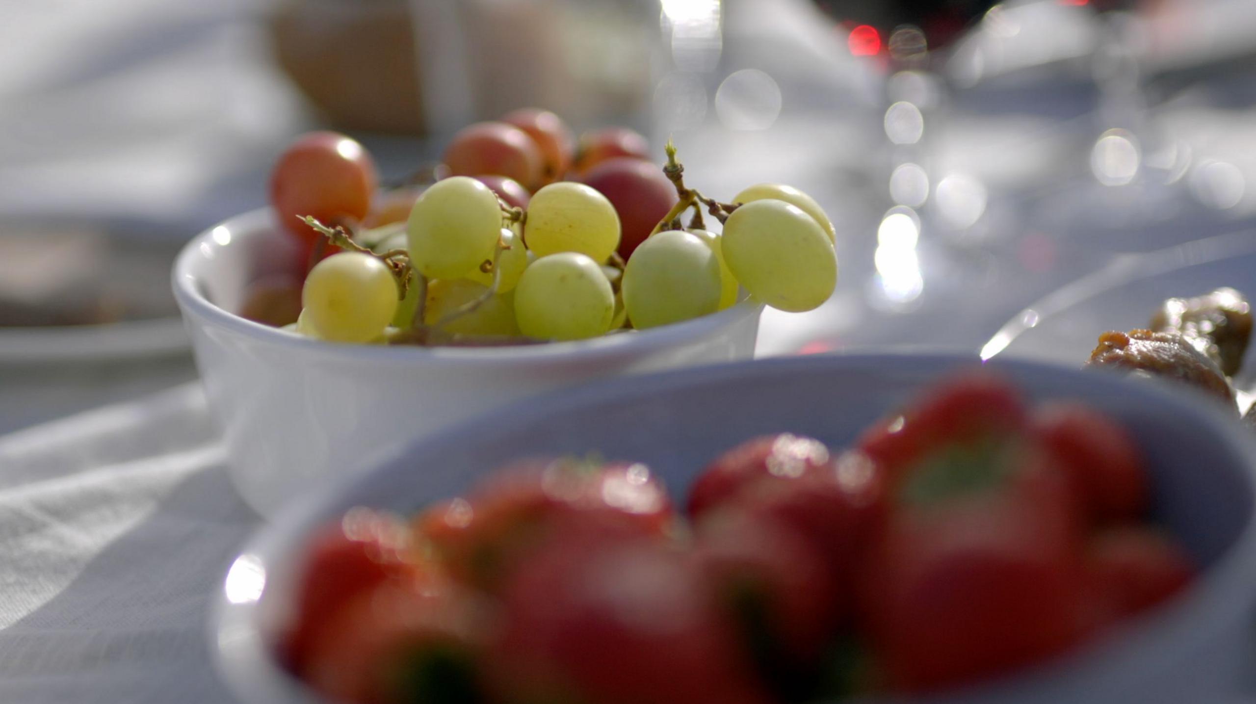 Close-up of a white bowl filled with green and red grapes on a table covered with a white cloth. In the foreground, another bowl contains red strawberries, slightly out of focus. The background shows blurred glassware and other picnic items, suggesting an outdoor dining setting in bright natural light.