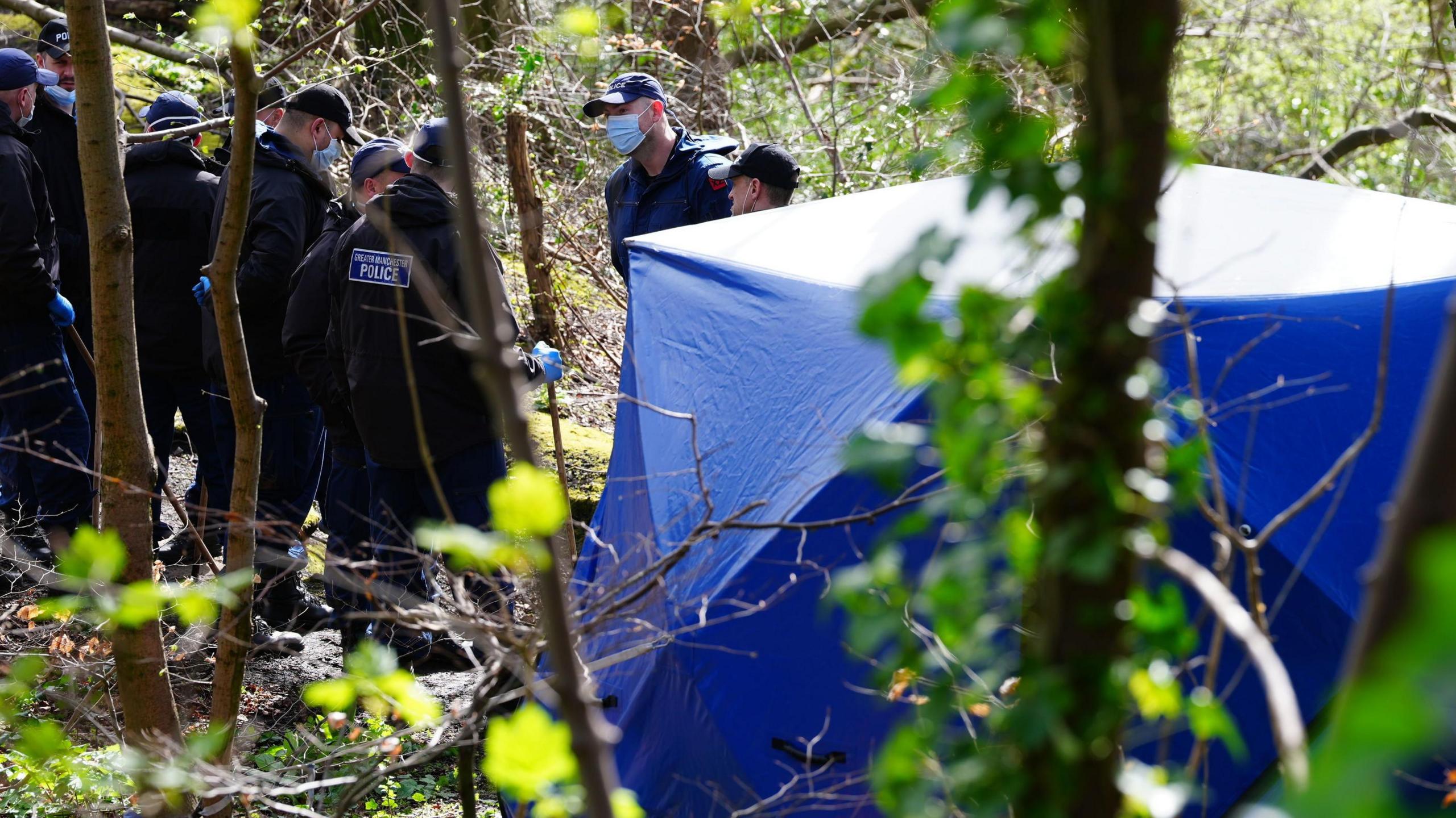 Police officers gather in the woods outside a blue tent - they are wearing face masks and caps and are in black uniforms with Greater Manchester Police on the back