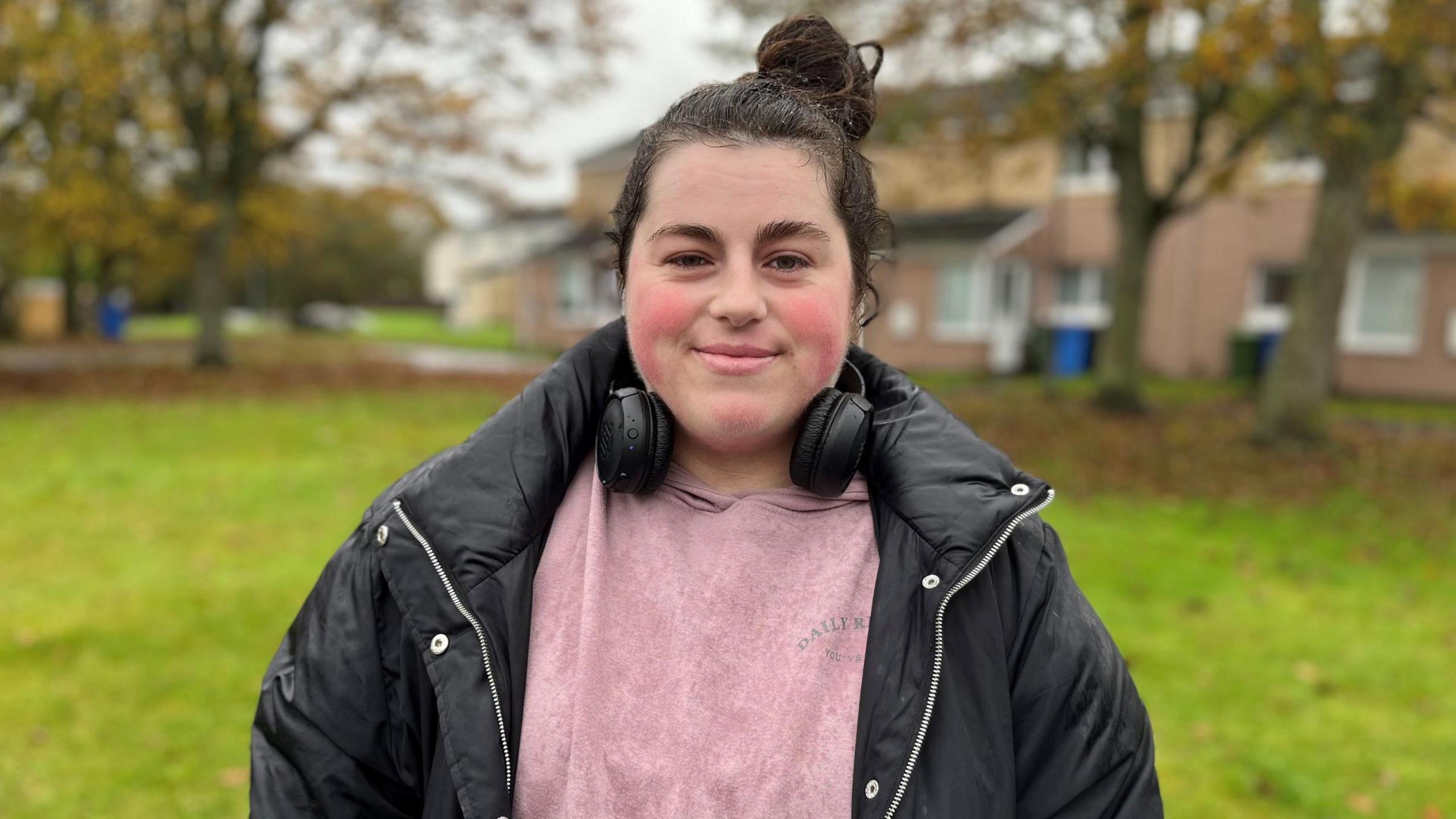 Courtney Wilson, a young woman, is standing outside. She has dark hair which is tied up and is wearing a black jacket over a pink top. She has a set of headphones around her neck. Behind her is a grassy open space and trees.