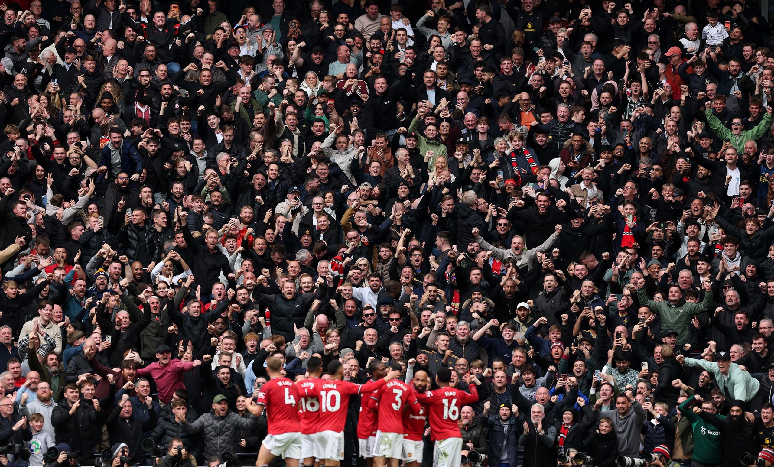 A group of football players in red jerseys stand together near the pitch, facing a large crowd of cheering fans in a stadium. The fans are densely packed, many raising their arms and wearing dark clothing, with some holding scarves.