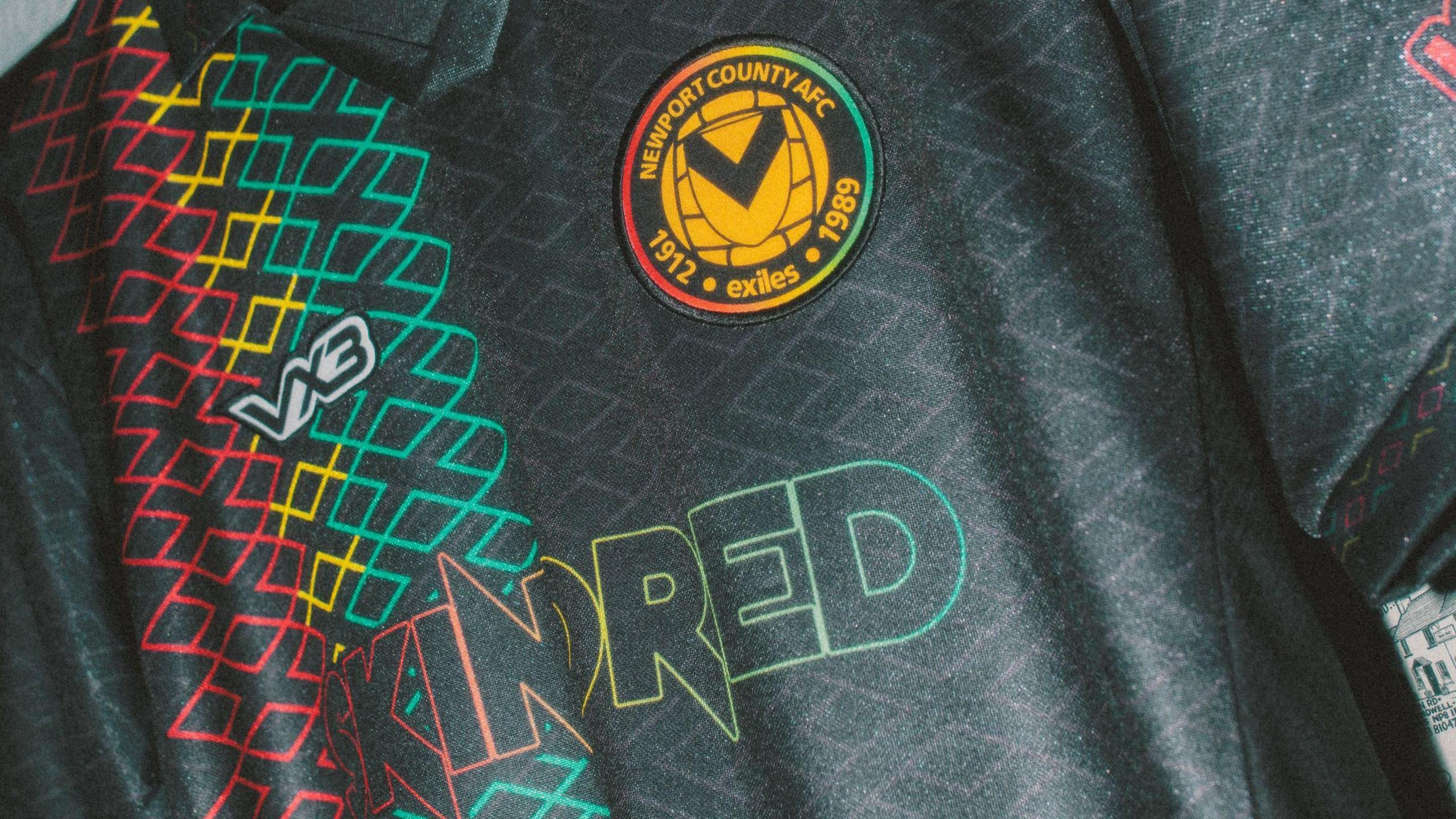 Close up shot of the black shirt, which features the stripes of the Caribbean colours in red, yellow and green and reads "Skindred" in their branding. The Newport County badge is yellow at the centre, with green, red and yellow around the rim. 