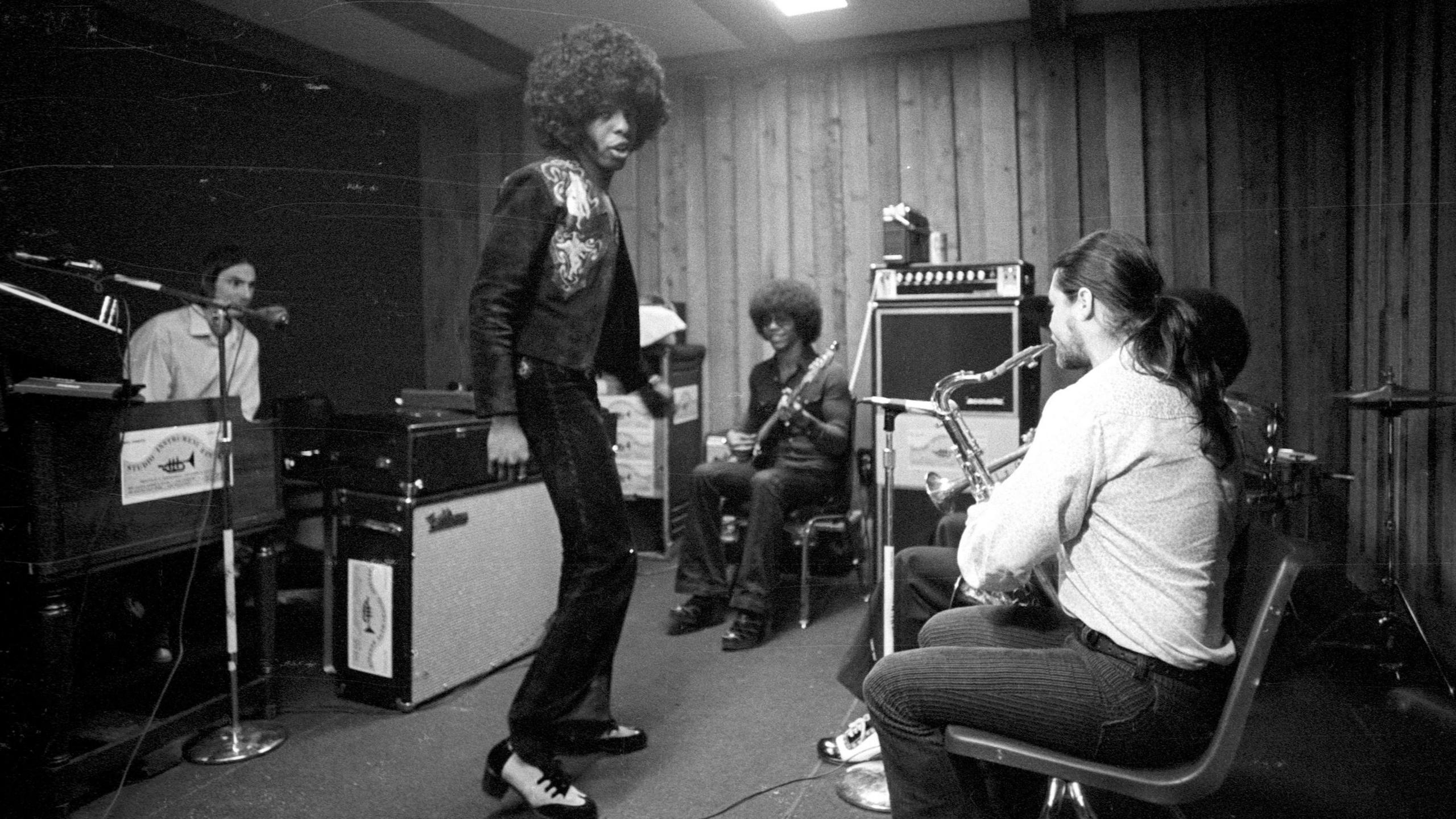Sly Stone sings and dances in the centre of a room with other members of the band pictured around him in a circle on various instruments. Two large amplifiers are dotted around the room.