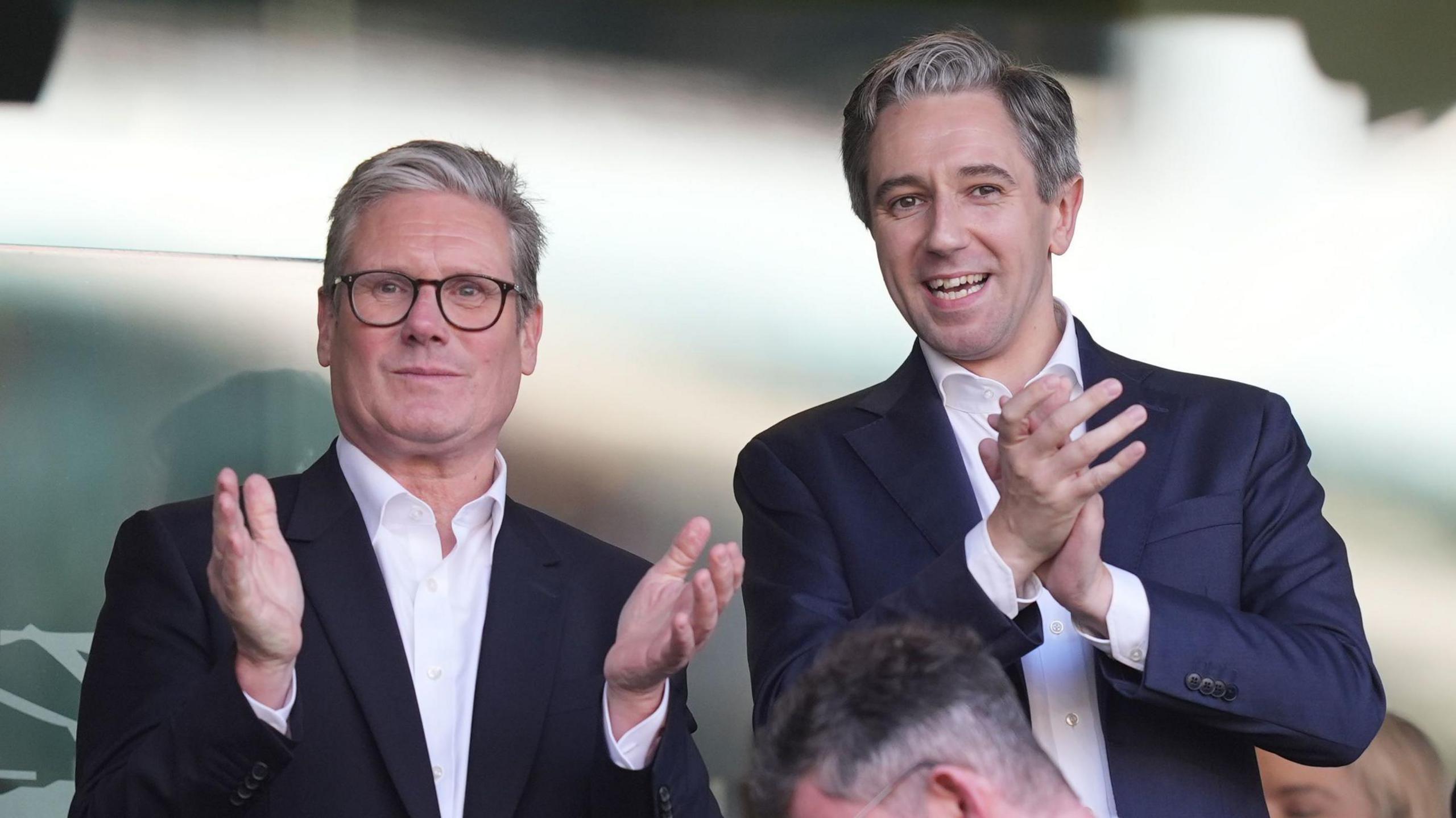 Sir Keir Starmer and Simon Harris, wearing dark suits and open neck shirts, standing and applauding in Dublin's Aviva stadium 