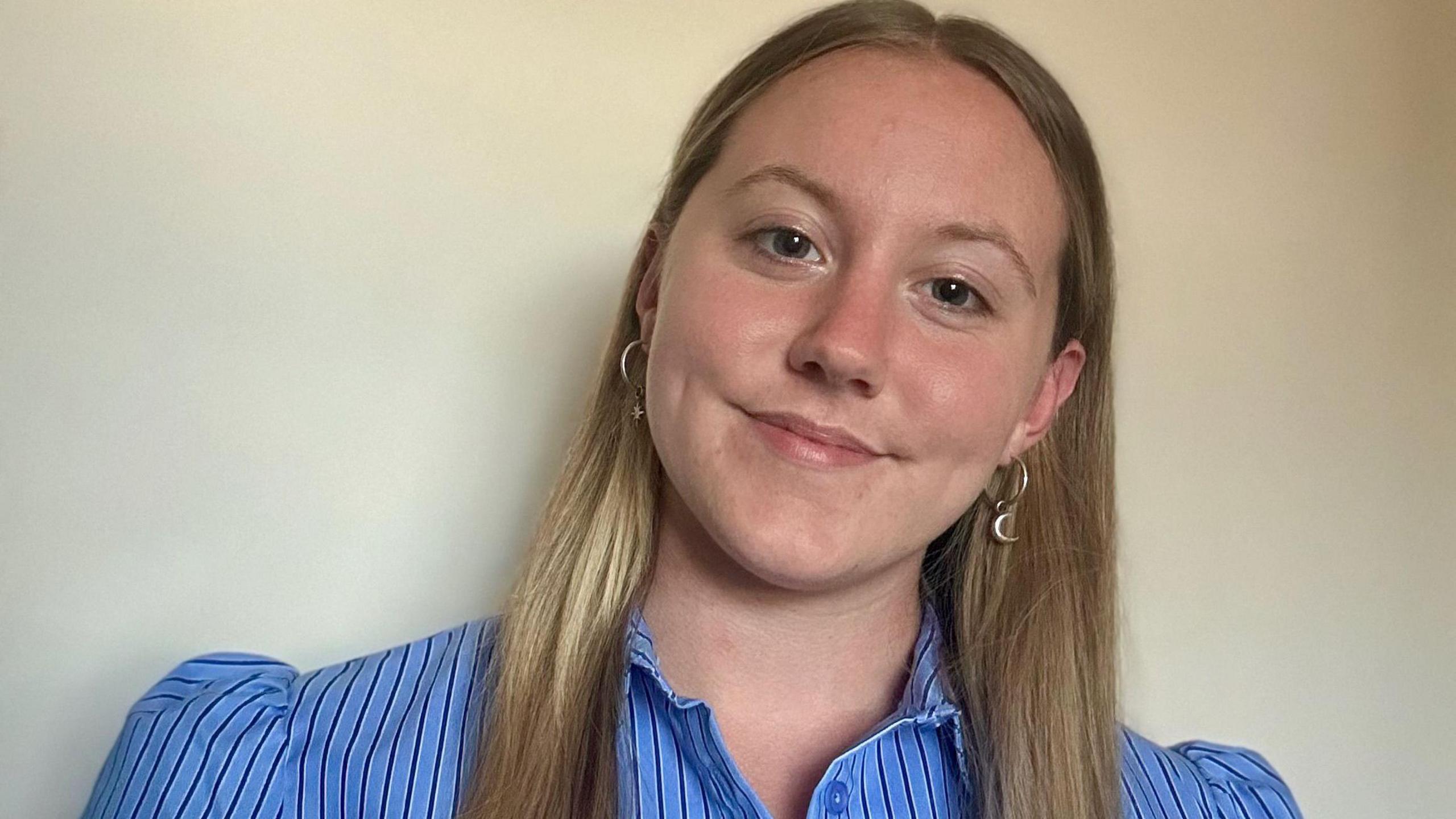 A woman with long blonde hair is wearing a striped blue blouse and smiling. There is a white wall behind her.