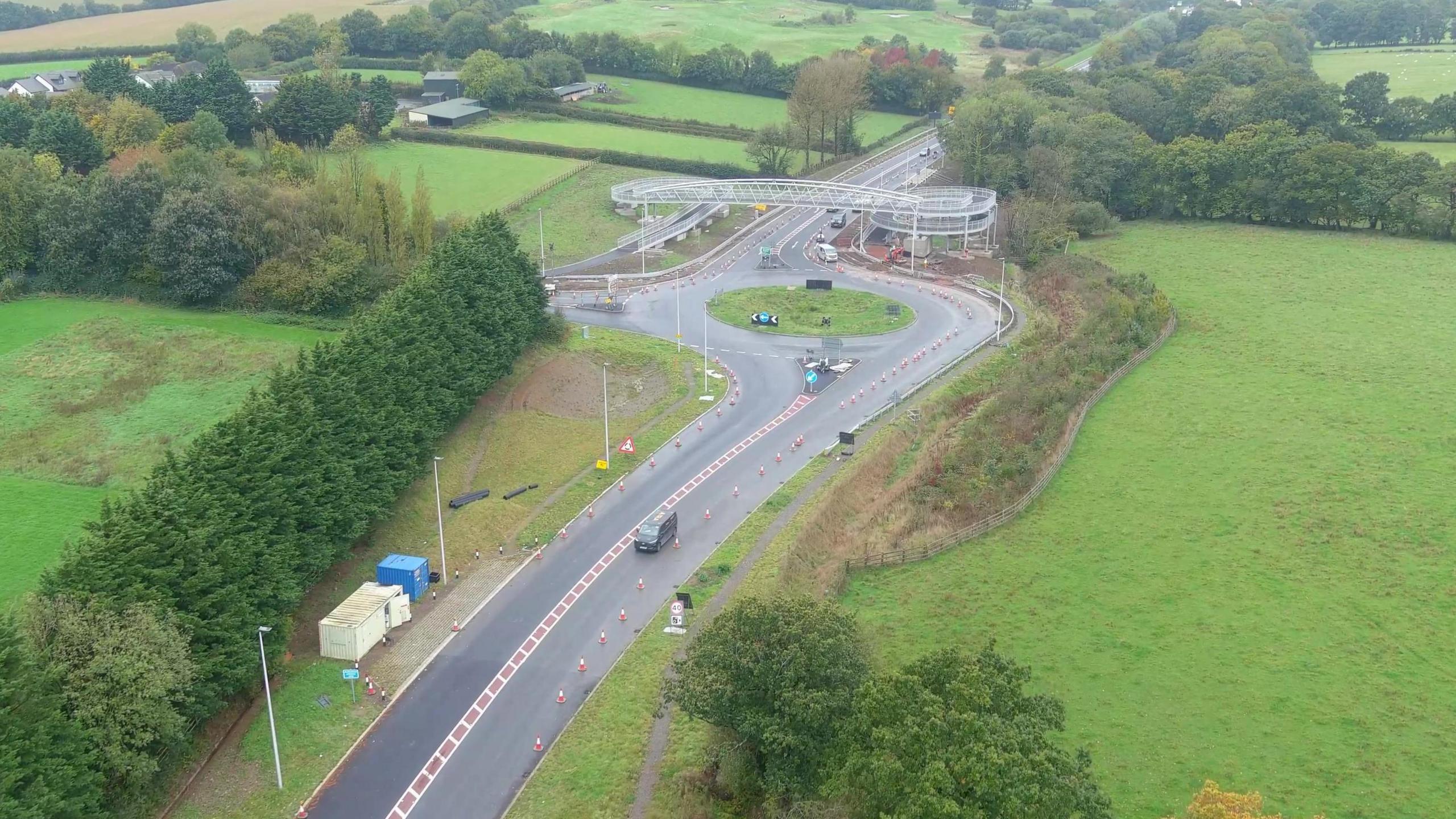 Part of the North Devon Link Road which has been undergoing roadworks. A set of traffic cones are on part of the road near a roundabout and a footbridge. The road is surrounded by fields and trees.