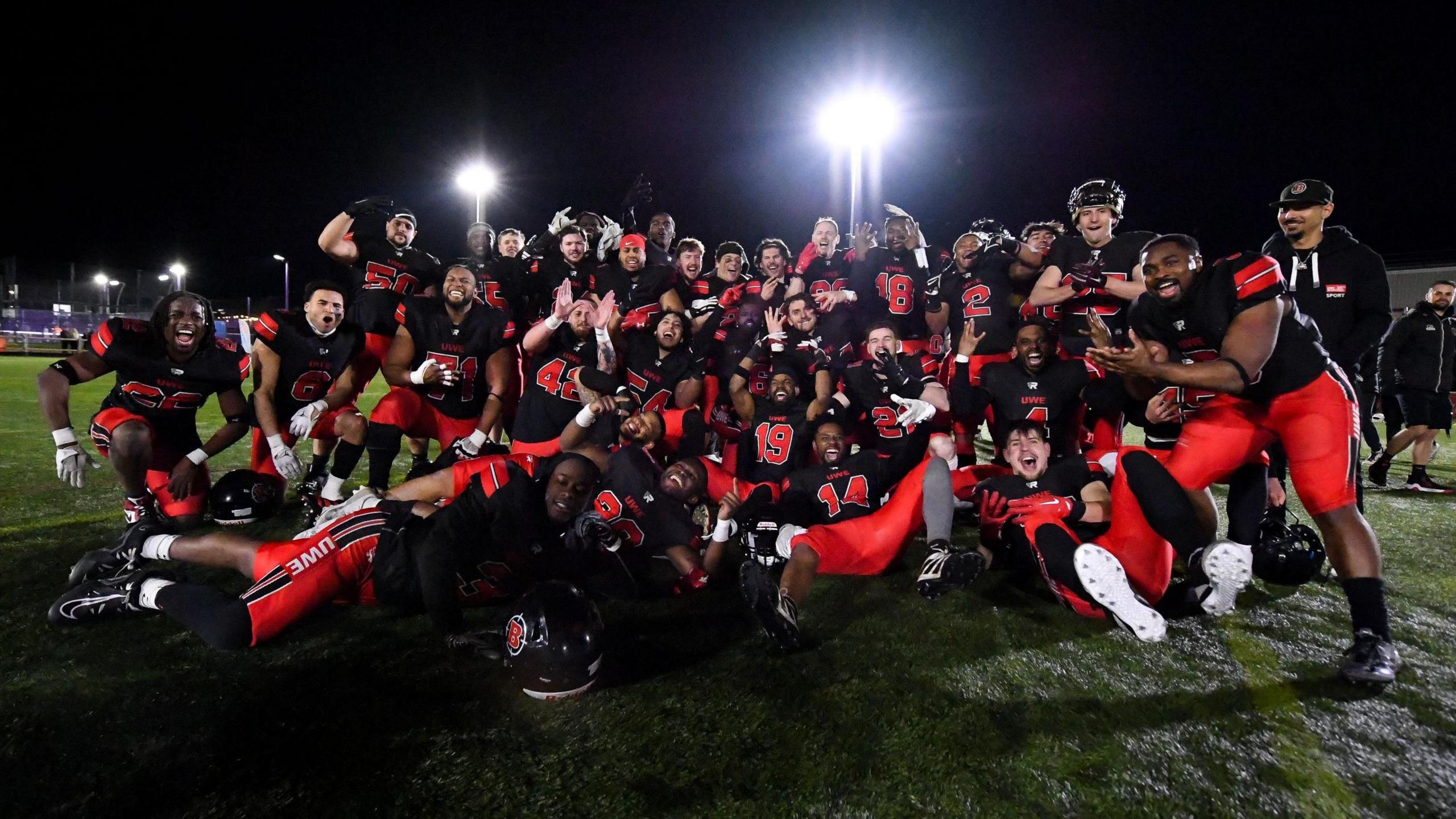 A large group of American Football players from UWE in Bristol celebrate winning a trophy. They are all in their kit of black jerseys and red trousers and are huddled together on a sports pitch illuminated by floodlights