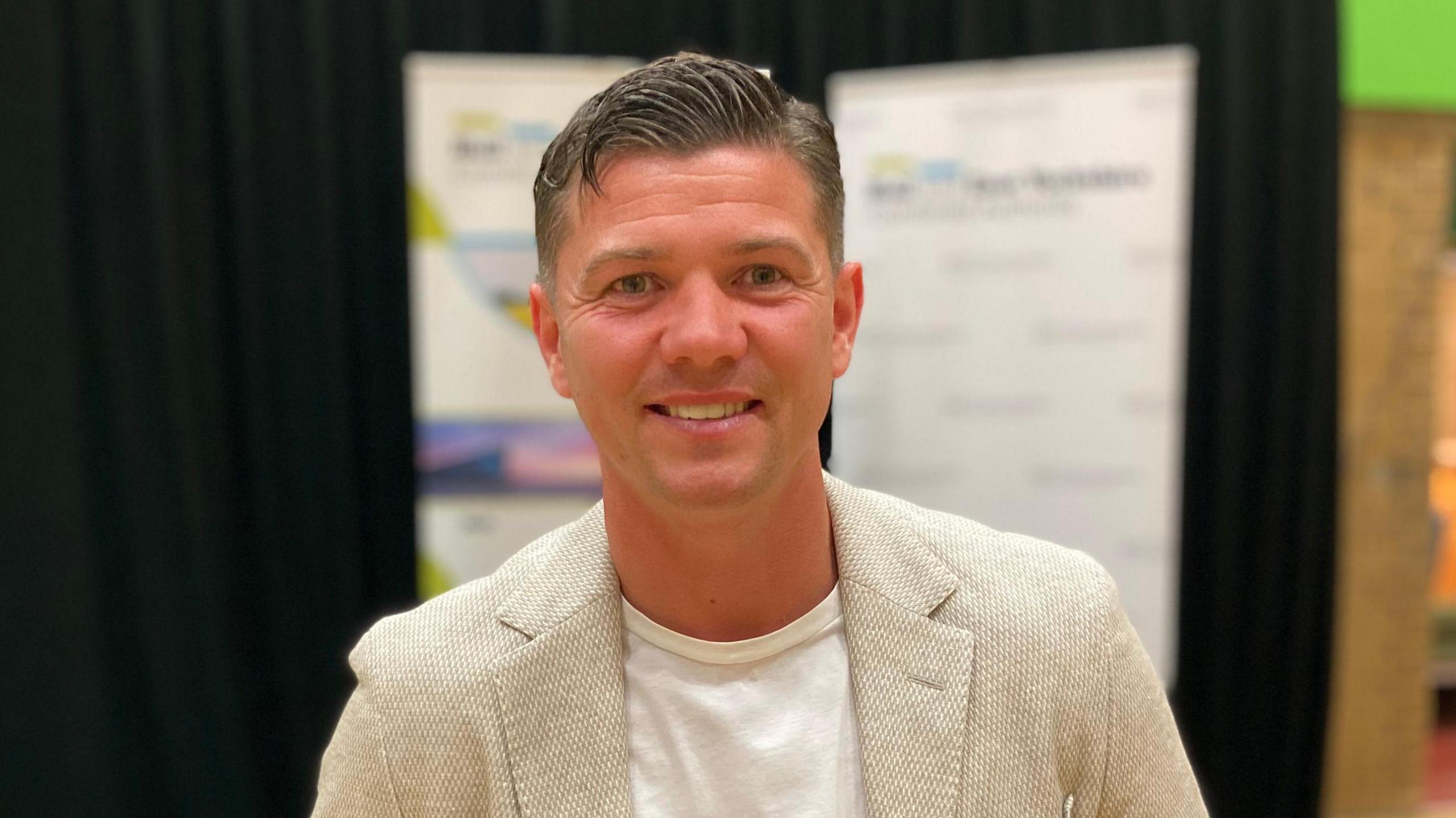 Luke Campbell smiles at the camera after winning the mayoral election in Hull and East Yorkshire. He is wearing a beige blazer with a white T-shirt. He is standing in front of a stage that includes a podium and a black backdrop.