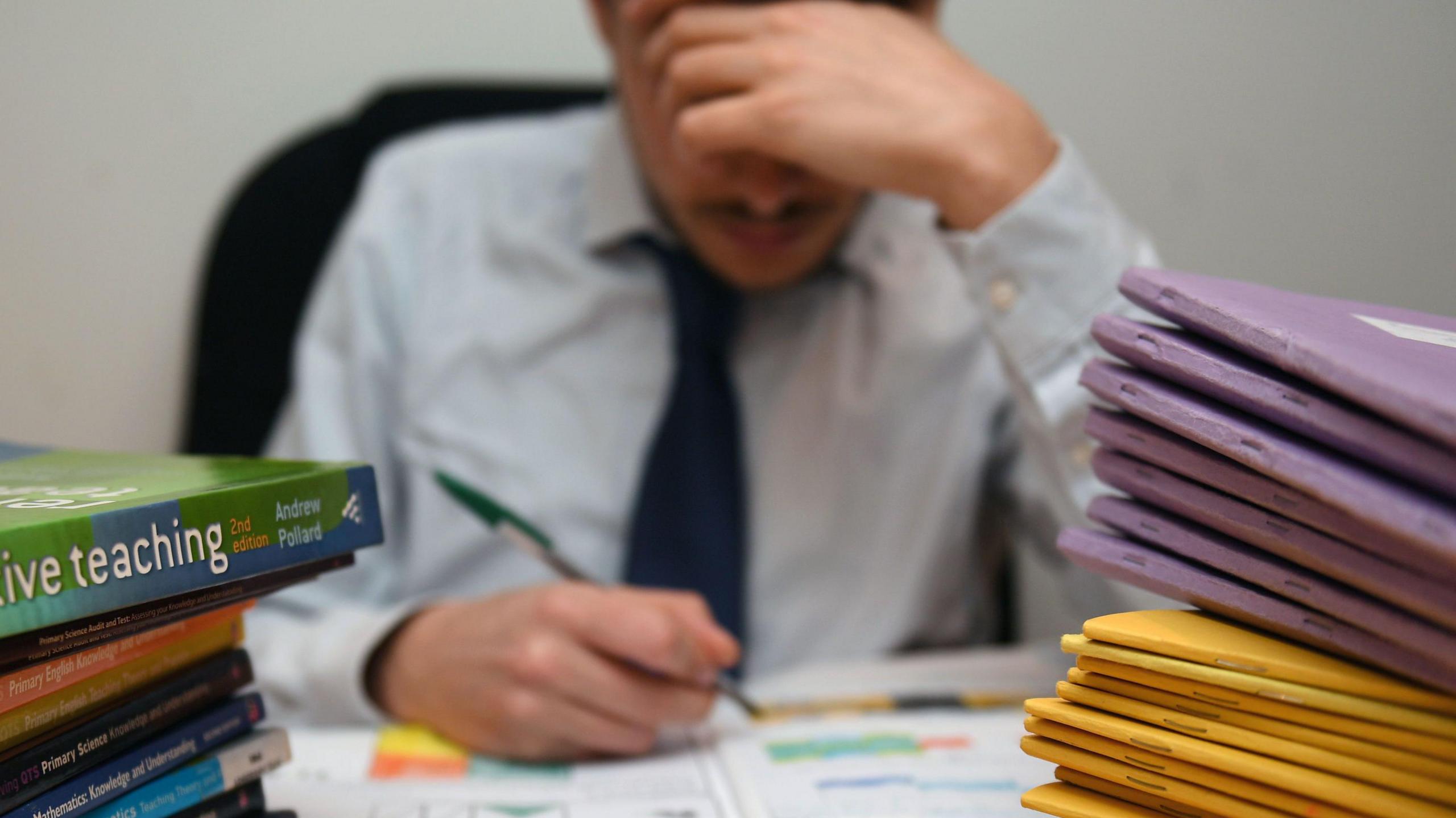 Photo of a school teacher looking stressed next to piles of classroom books.