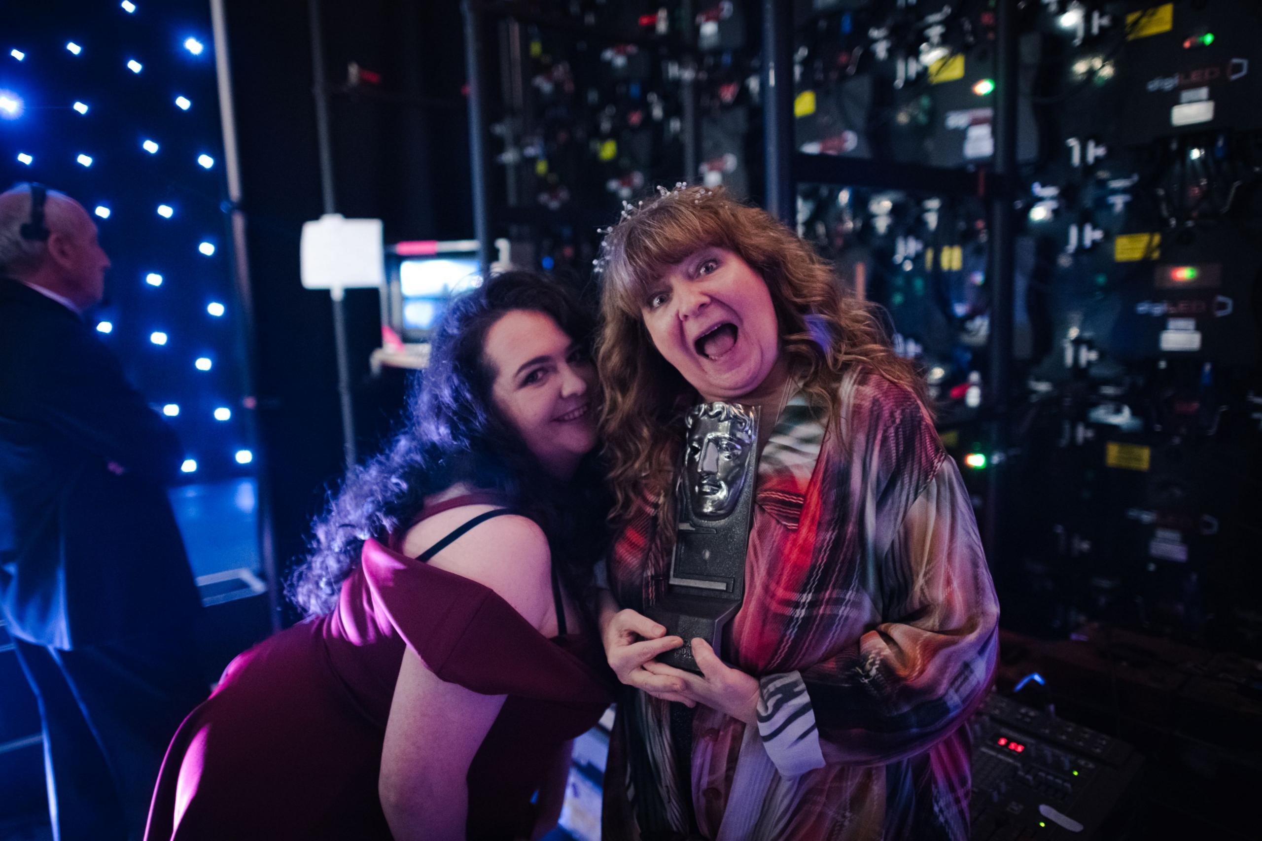 Backstage at a previous Bafta ceremony, Janey Godley, in a tartan chiffon gown and tiara, holds her own Bafta award while posing with her daughter Ashley, who leans in to her mum and wears a plum-coloured strappy evening gown.