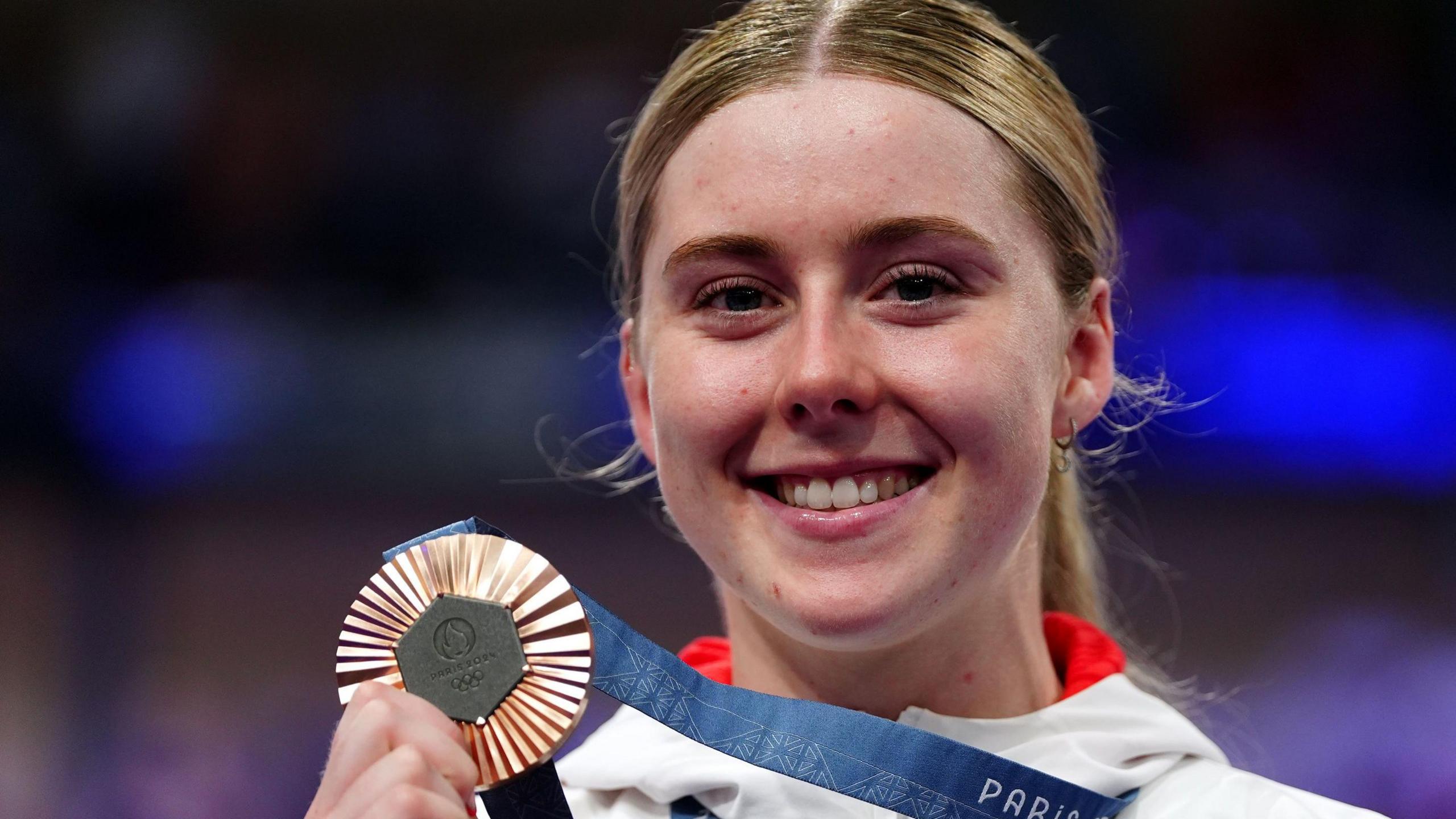 A female cyclist smiles as she holds up an Olympic bronze medal attached to a blue ribbon, which she is wearing around her neck. She has blond hair, tied back, and is wearing a white tracksuit top with red trim.