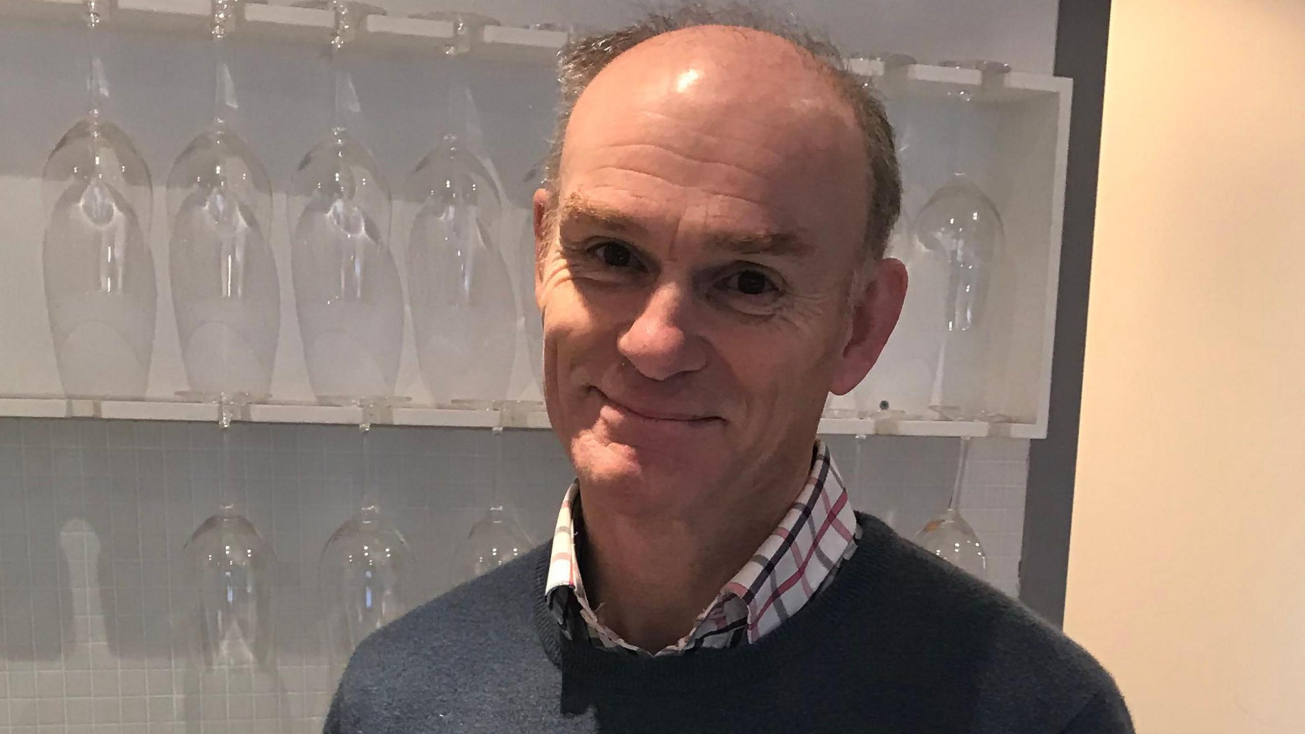 Philip Tuck in a blue jumper with a checked shirt, standing in front of shelves with wine glasses on.