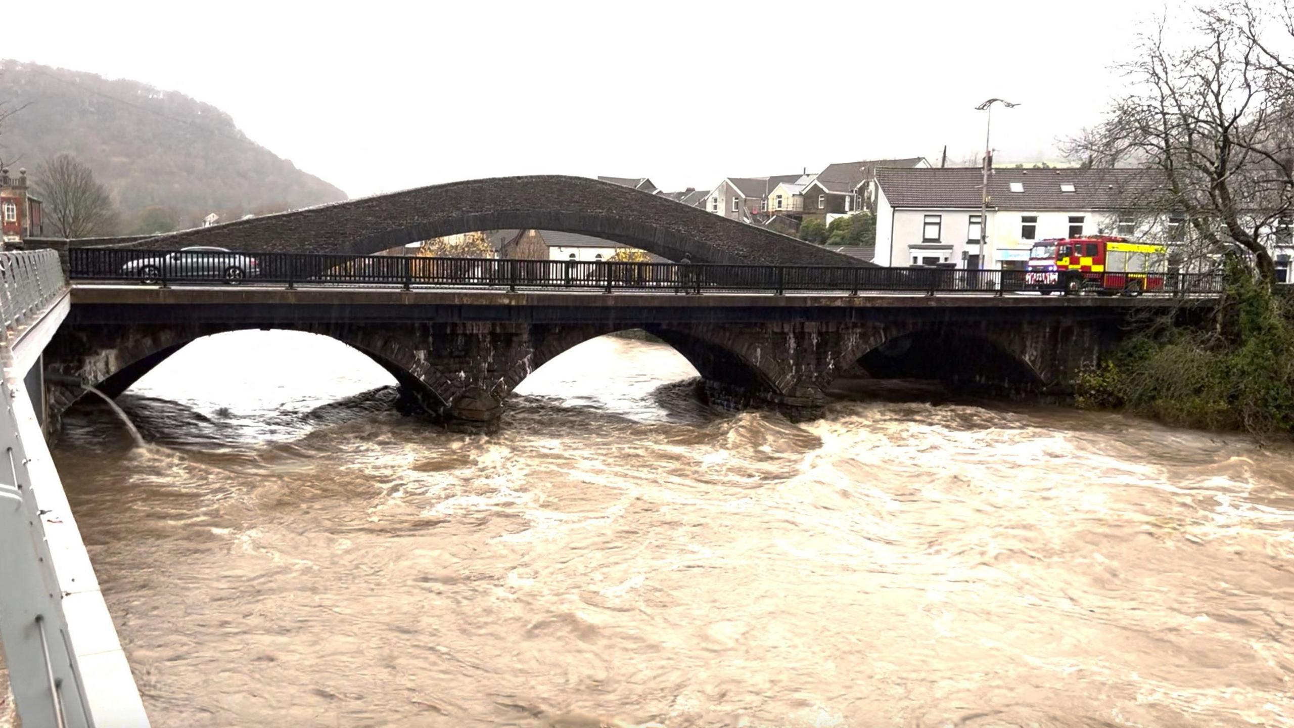 A swollen river runs under a road bridge, with a car and fire engine on the bridge and houses in the background.