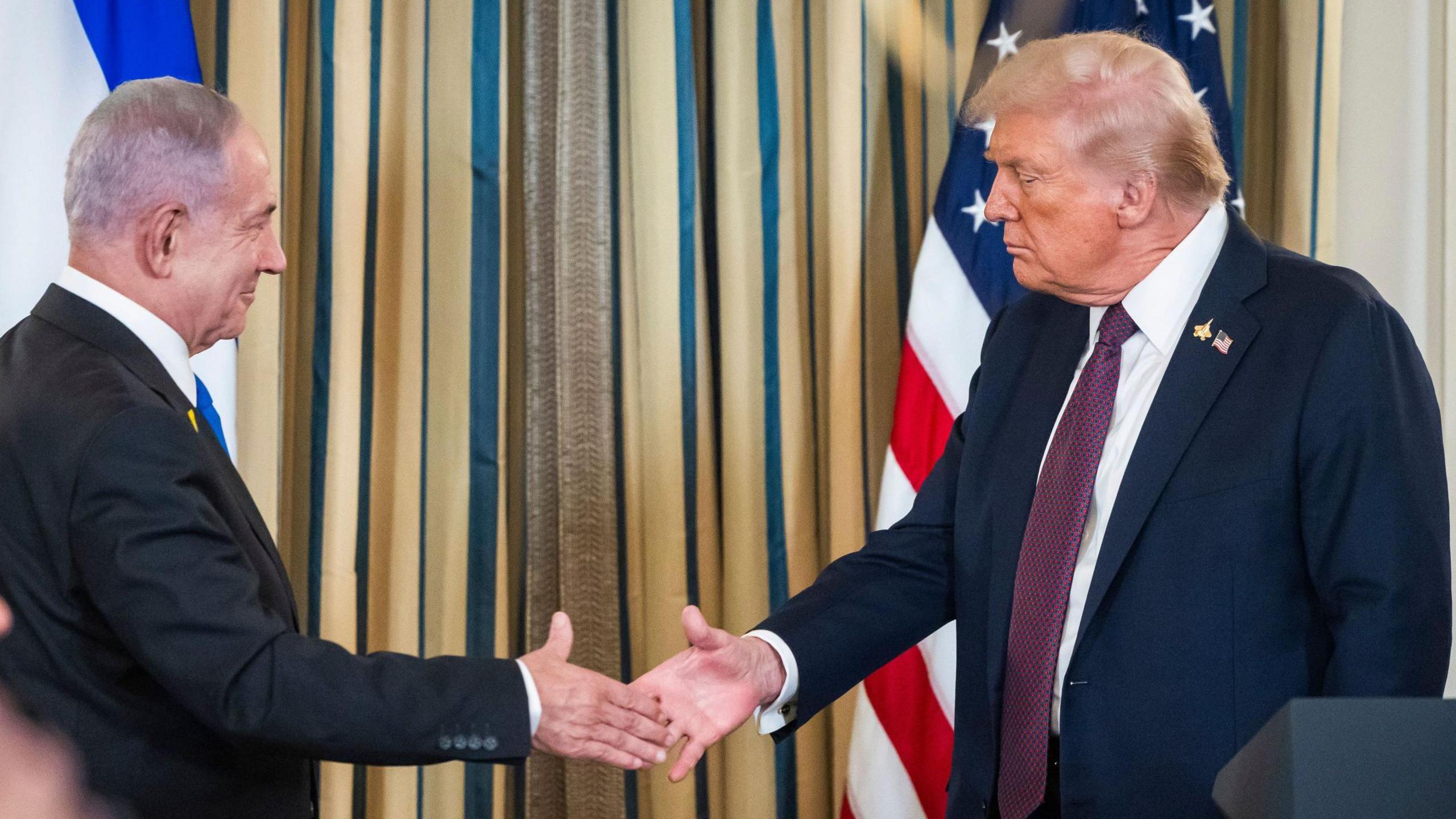 US President Donald Trump (R) shakes hands with Israeli Prime Minister Benjamin Netanyahu (L), with Israeli and American flags behind them. Both men are wearing dark suits and white shirts.  