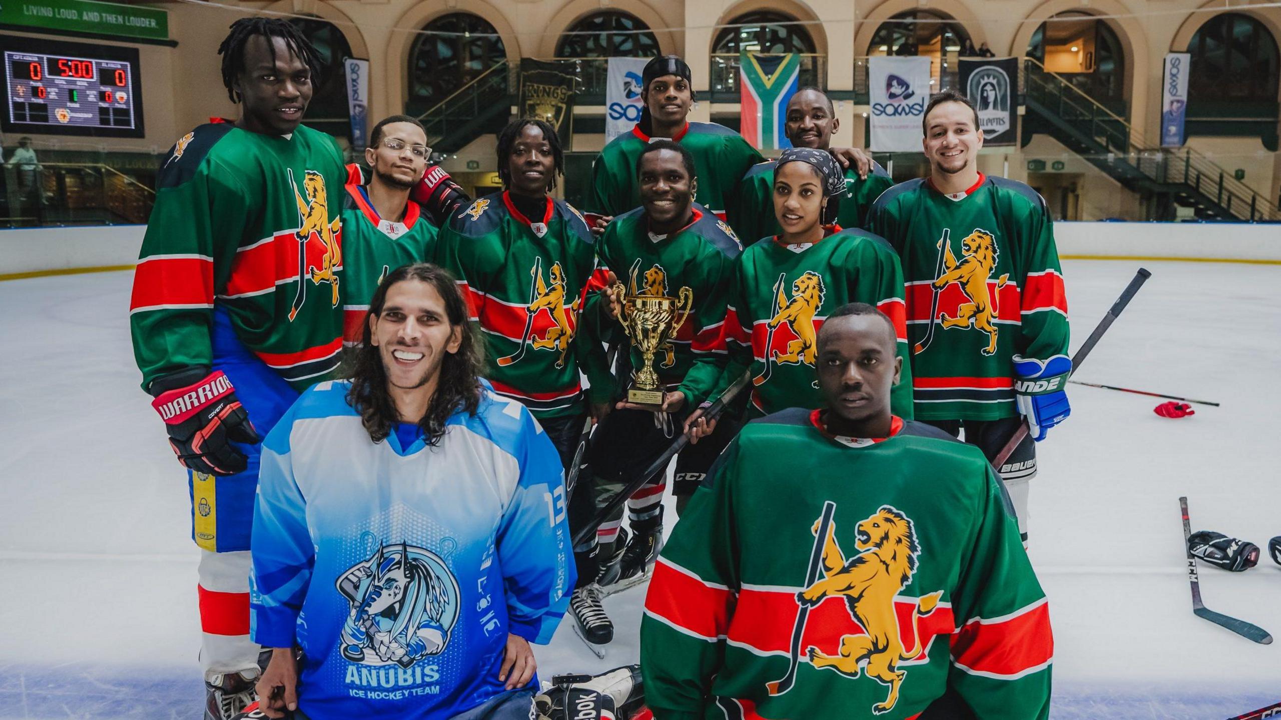 A group of 10 Kenya Ice Lions players, mostly wearing green and red striped ice hockey jerseys, pose for a team photo while standing on an ice rink and holding a gold trophy