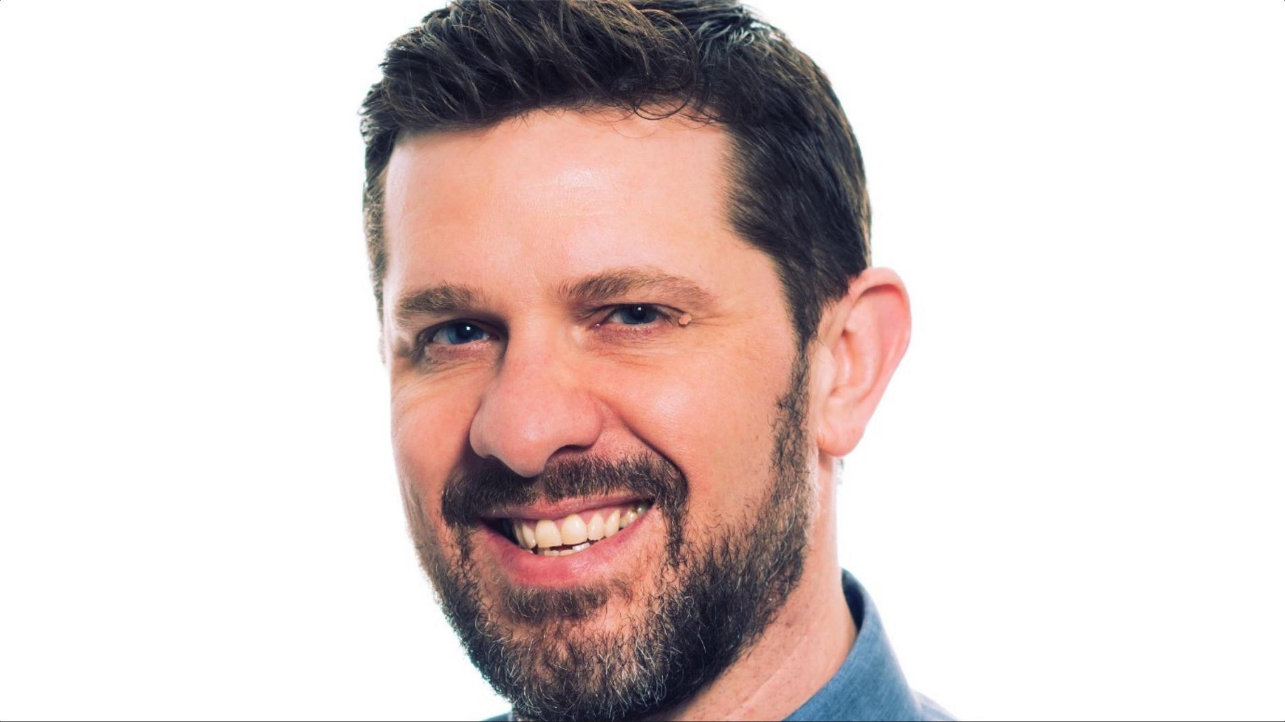 Kevin Mapstone with dark hair and beard, smiling and wearing a blue shirt against a white background