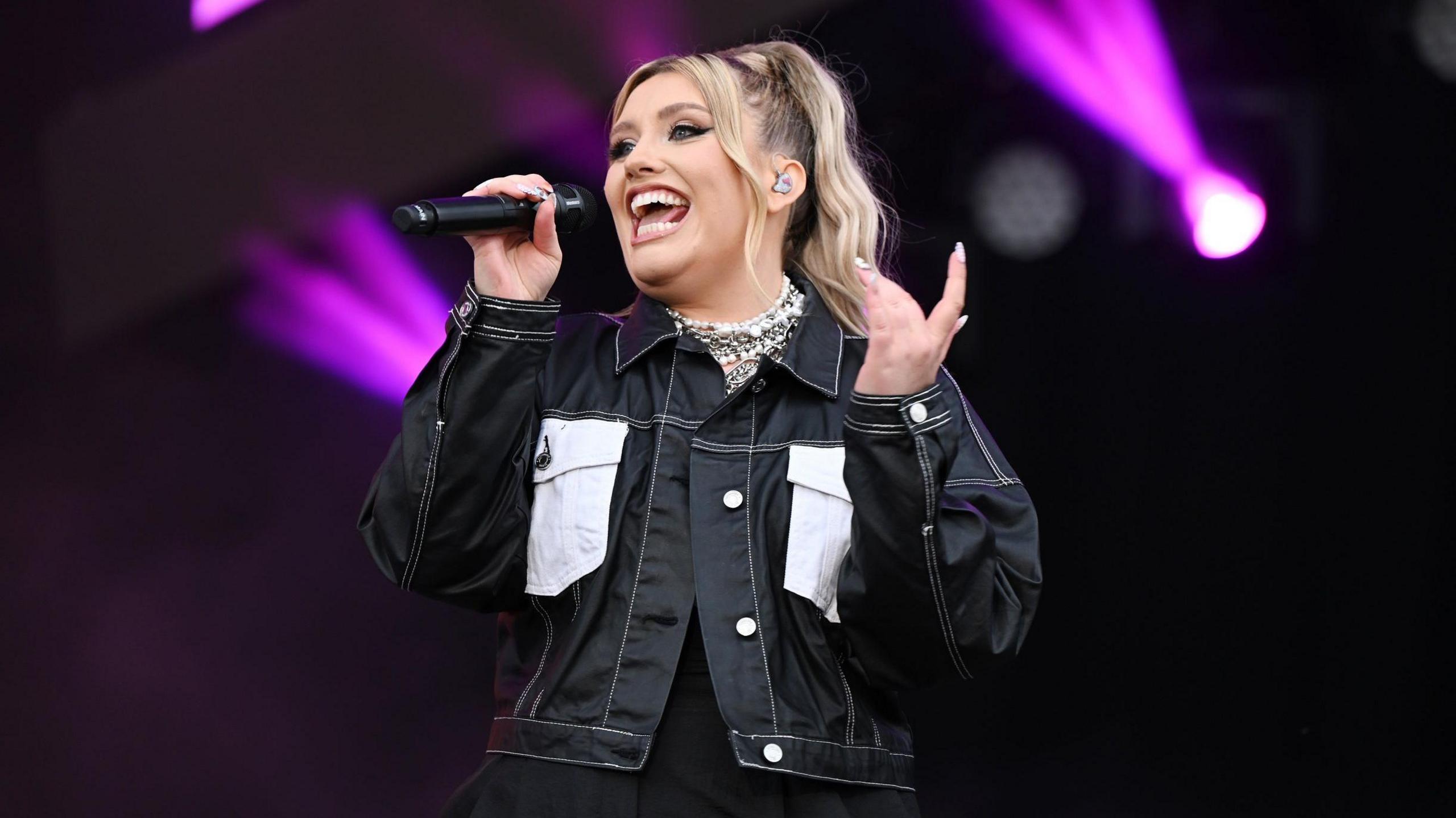 Ella Henderson on stage singing into a microphone wearing a black jacket with her hair in a ponytail