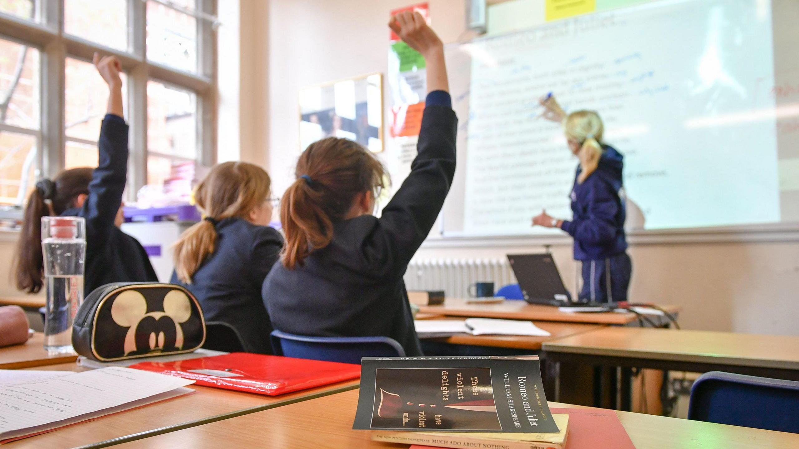 Anonymised image of pupils in school classroom with hands raised, as a teacher points to words on a whiteboard.