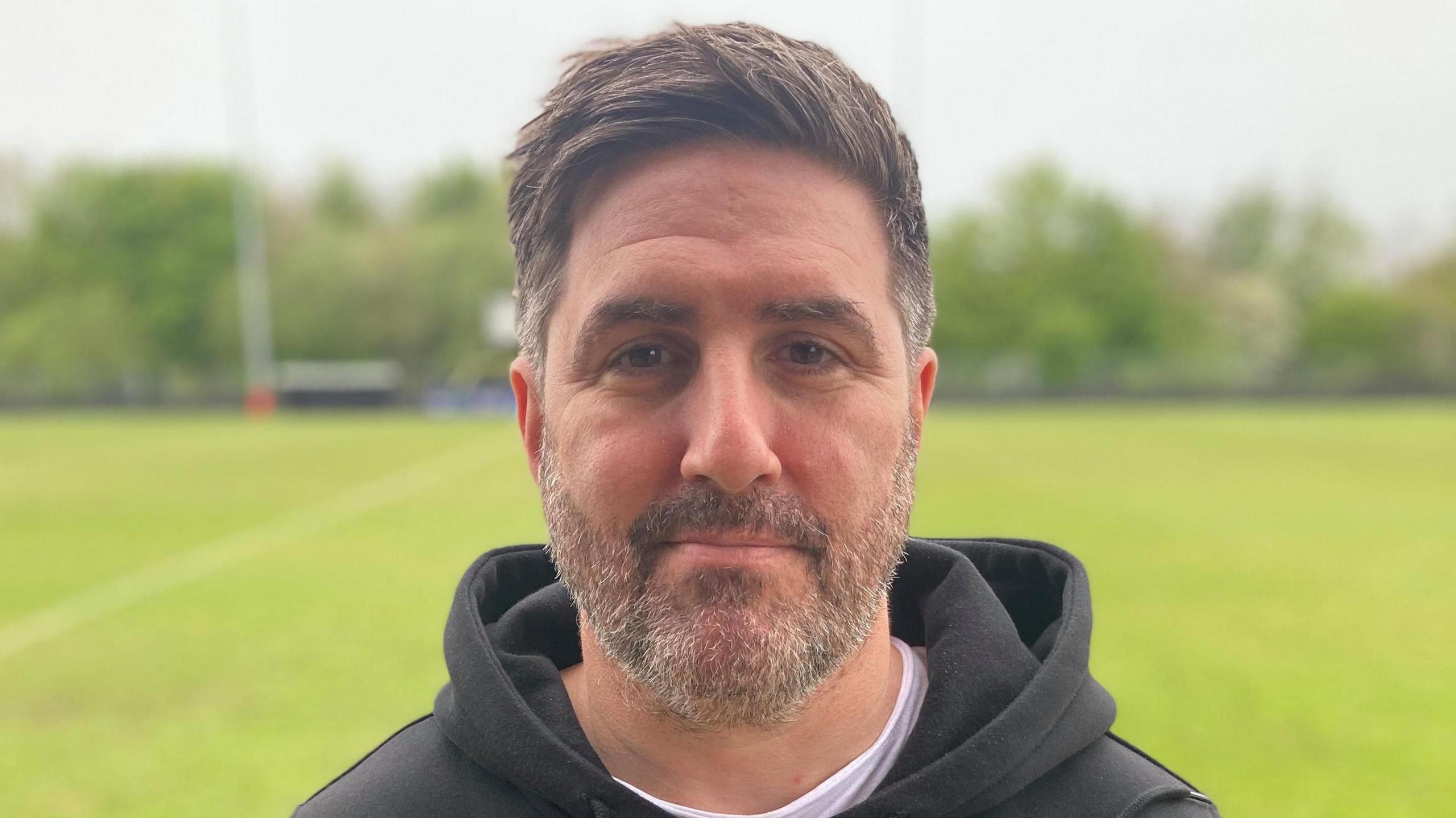 A man with brown hair and short beard wearing a brown hooded top standing on a football pitch. 