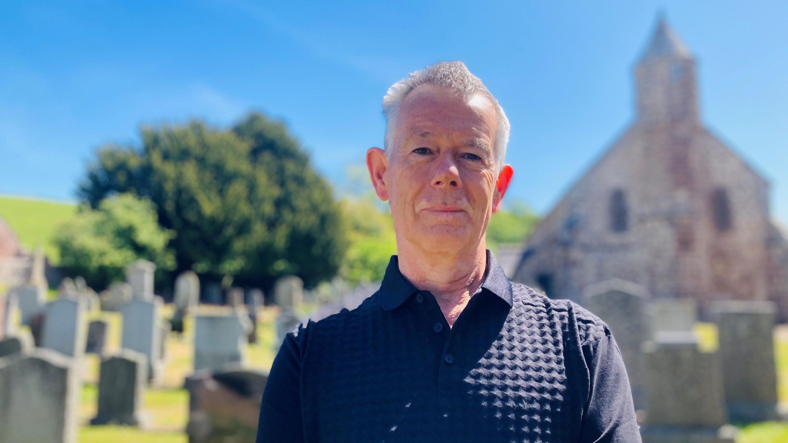 Man - Charles Roberts-McIntosh - standing in graveyard, with Arbuthnott Church in background, under a blue sky.