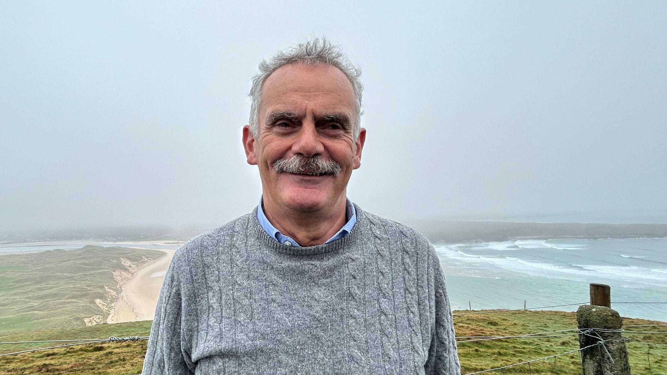 Andrew Ward, a man with short grey hair and a grey moustache, stands on high ground with a beach and stormy sea in the background. He is wearing a grey cable-knit sweater with a blue shirt underneath.