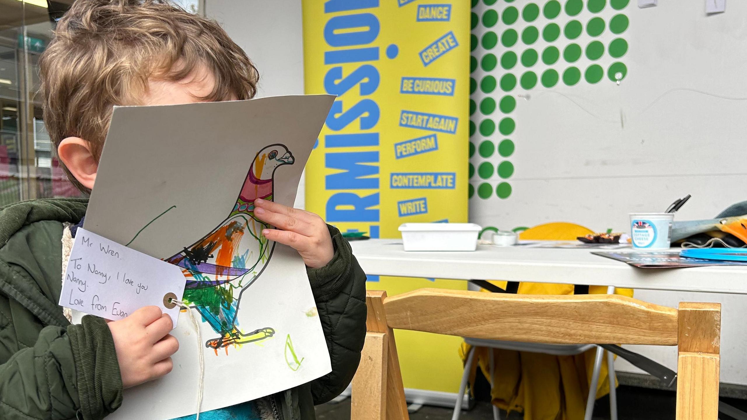 A young boy in a green coat covers his face with his design and a note to his nanny. A work table with art supplies and a wooden chair are in the background.