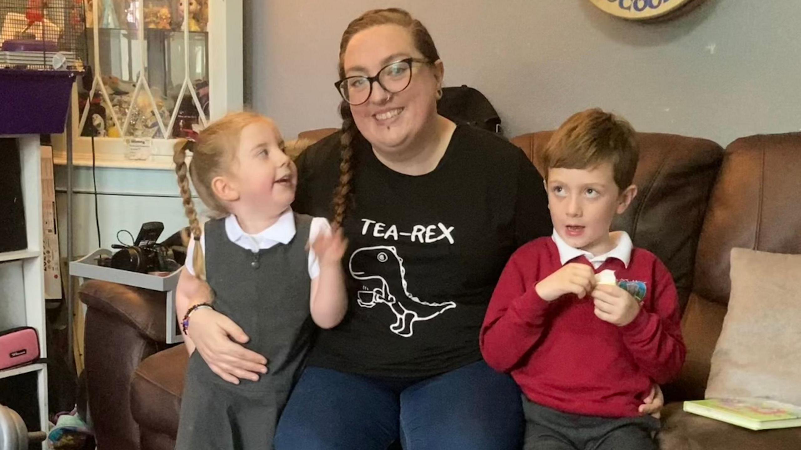 Debbie McCrudden and her two children sitting on a sofa. Conall to the right wearing a red school jumper, and Lilly to the left wearing a grey school dress.