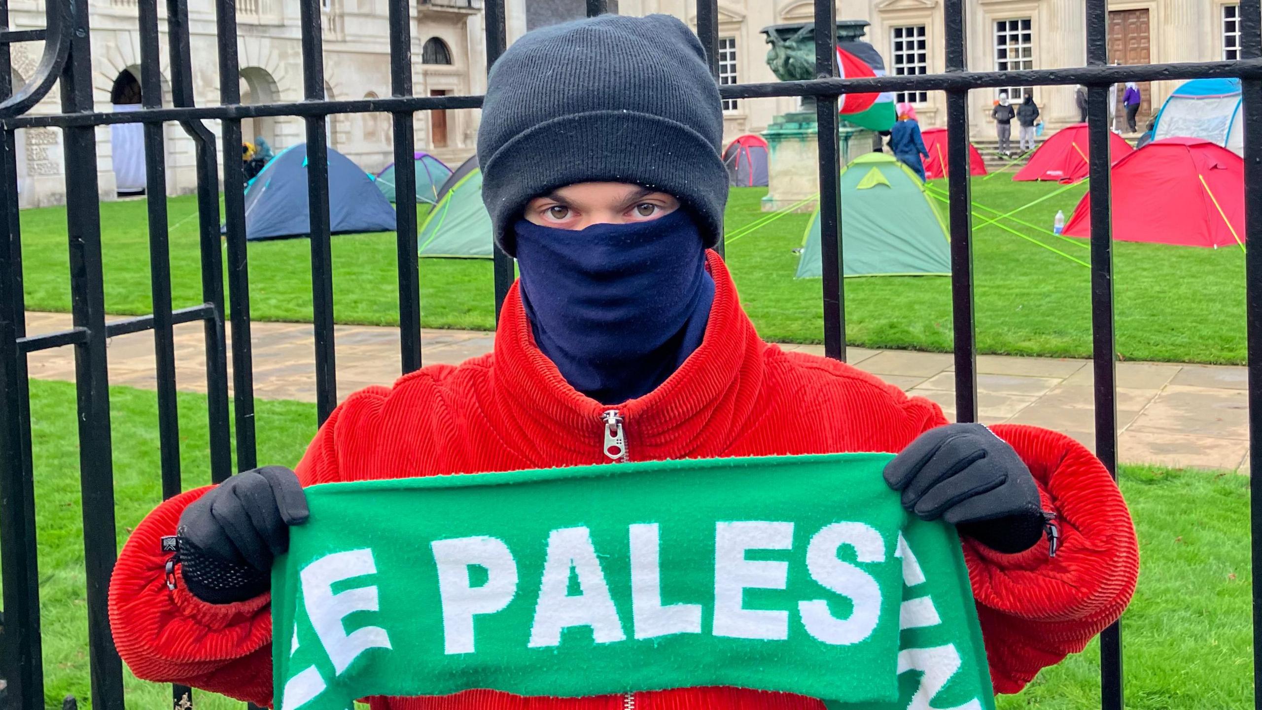 Morris is wearing a red zip up corduoroy coat, with black gloves, a blue face covering and a black hat. He is holding a green banner which says PALESTINE on it. He is stood in front of railings outside the front of Senate House. Numerous tents can also be seen on the lawn behind him.