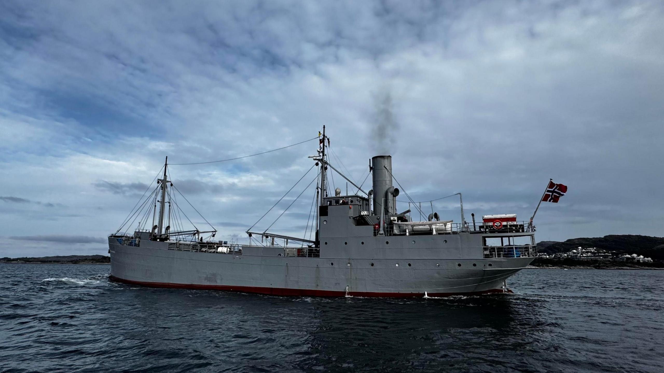 View of 200ft (60m) steam ship - S/S Hestmanden. Grey metal, with a funnel, it has two derricks, or lifting devices and is flying the Norwegian flag from its stern.