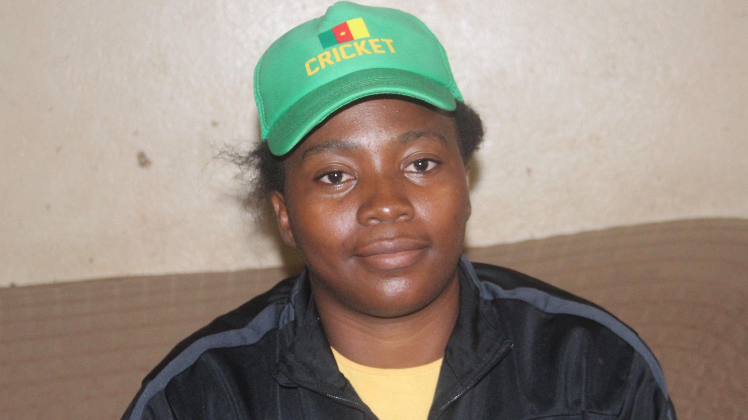 Madeleine Nseke Sissako looks at the camera while wearing a green baseball cap bearing Cameroon's flag and the word cricket in gold stitching. She wears a black tracksuit top over a yellow t-shirt while sitting on a beige sofa