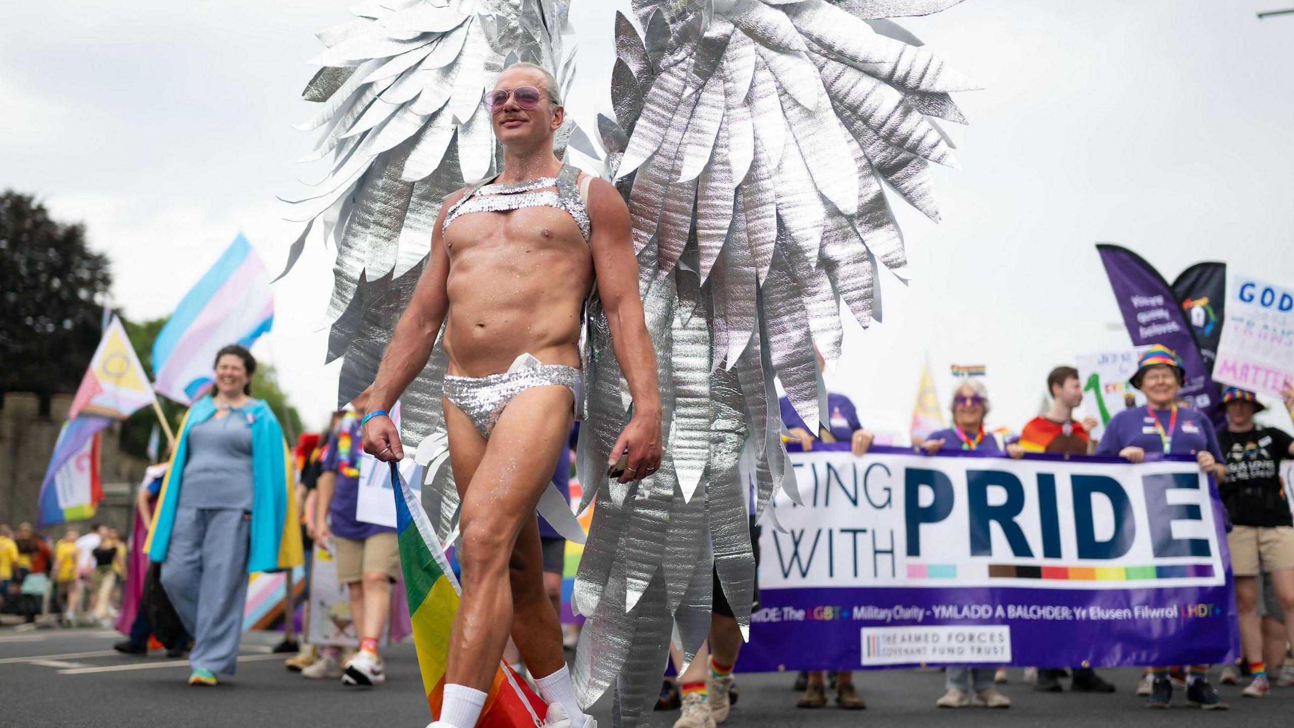 A man wearing silver thong has wings made of silver strips on his back as he walks in front of a crowd who are holding flags and banners