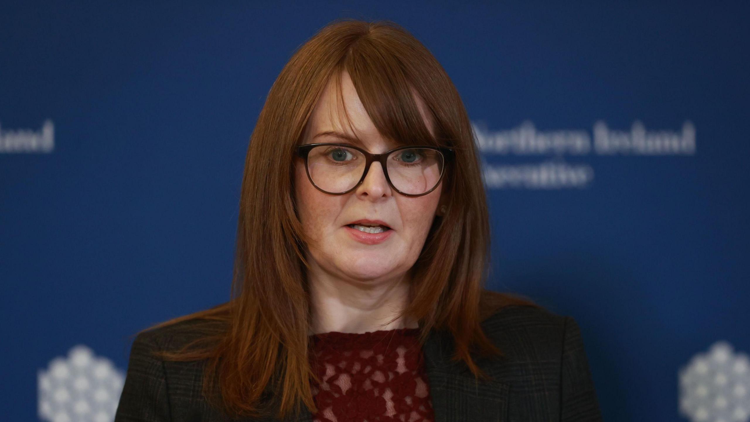 Caoimhe Archibald with long ginger hair, she is wearing glasses, a black blazer and a burgundy top.