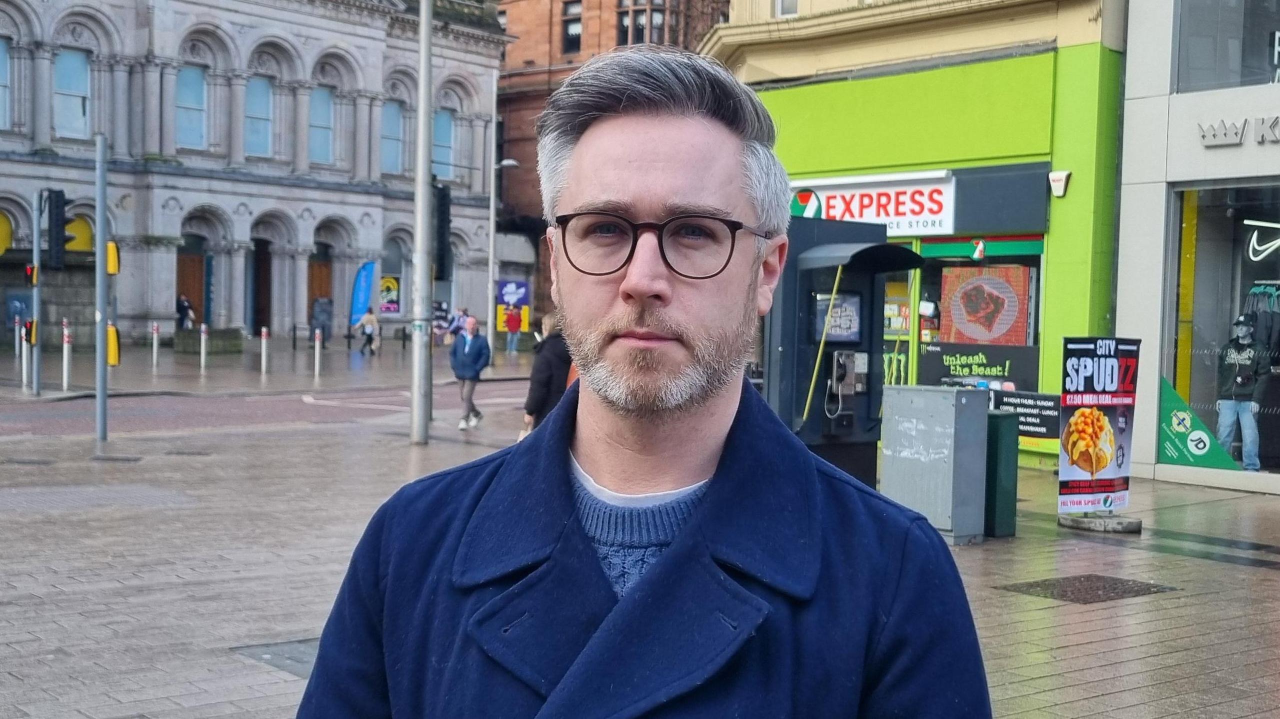 Man wearing glasses, a navy coast and light blue knitted jumper. He is standing in front of a telephone box and a fluorescent green convenience shop. 