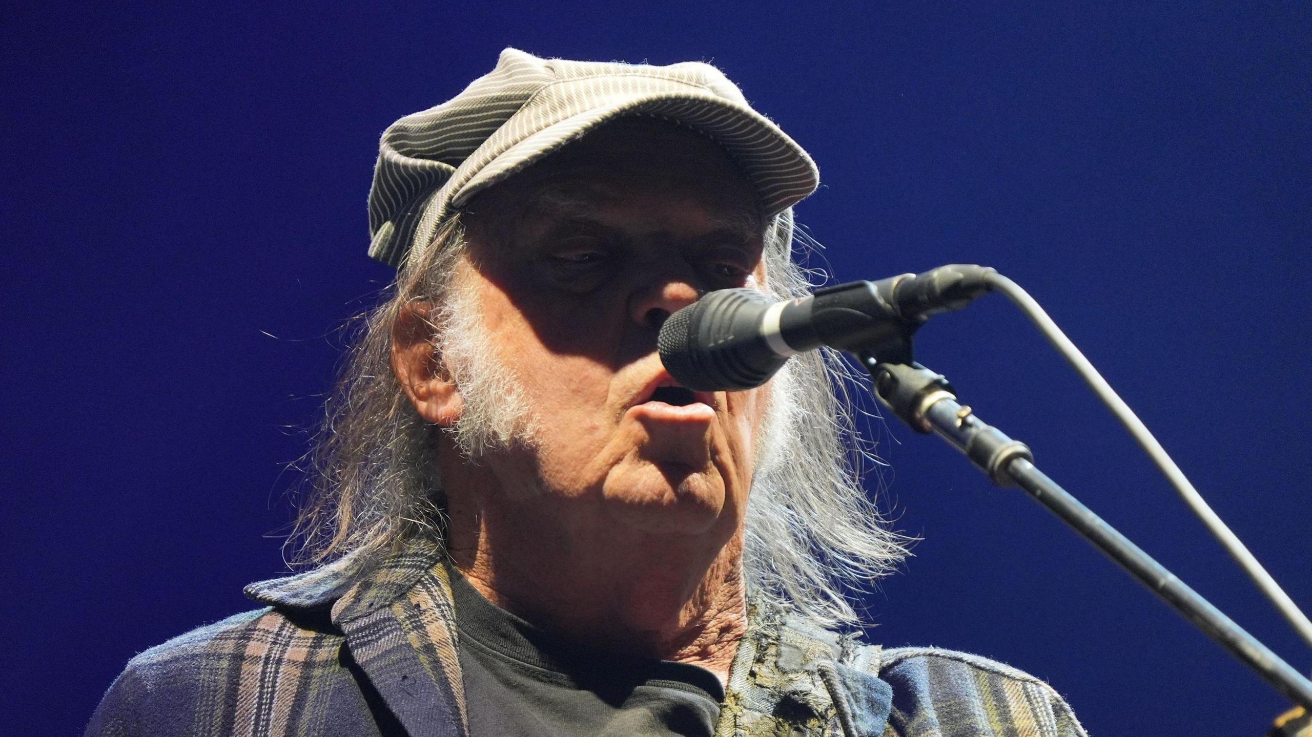 Neil Young sings into a microphone. He has long grey hair and sideburns. he is wearing a chequered navy jacket and a tan cap.