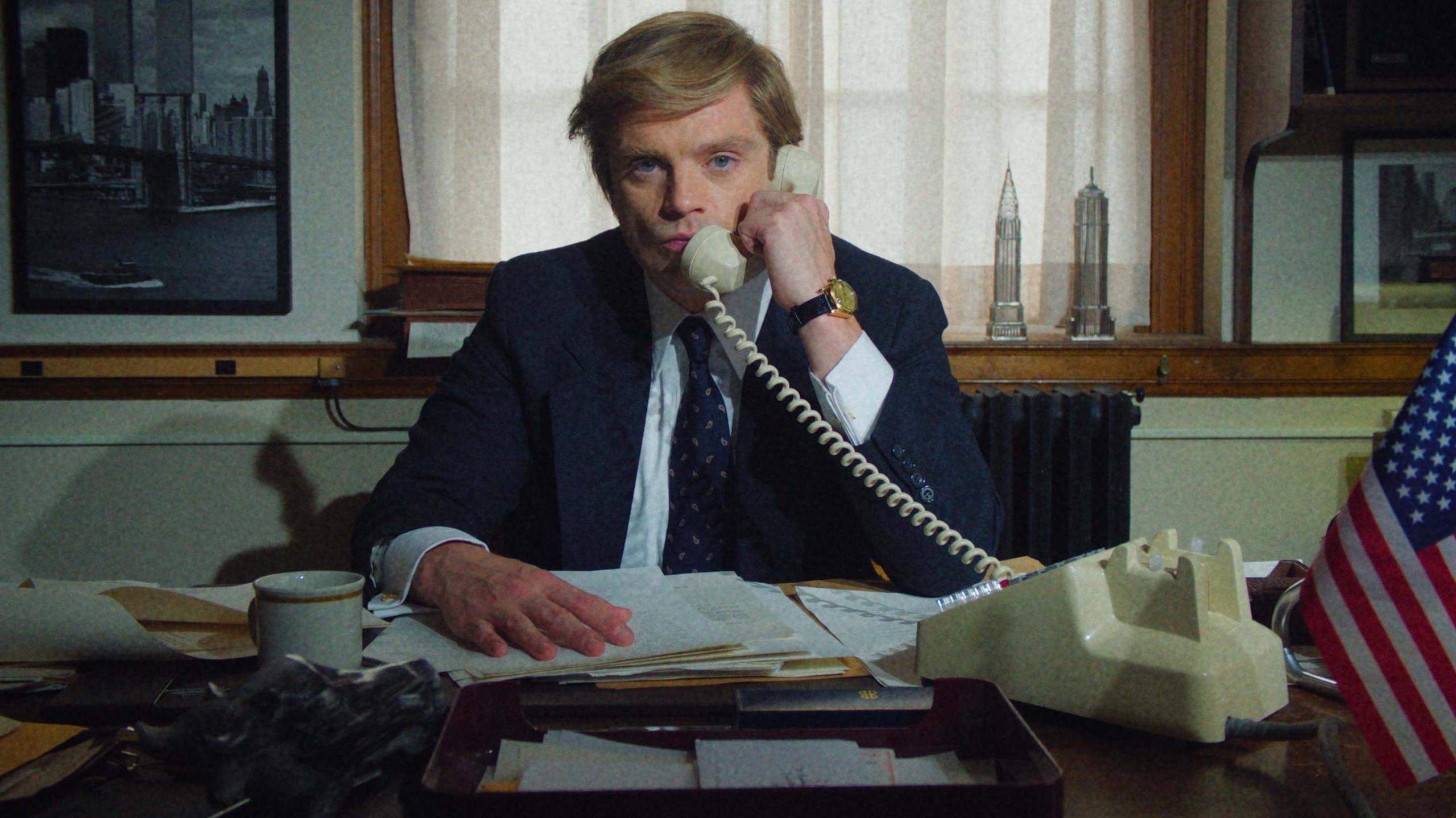 Sebastian Stan as Donald Trump in The Apprentice - he is on a white landline phone while sitting at a desk strewn with business papers. He is dressed in a navy suit and looks very serious.