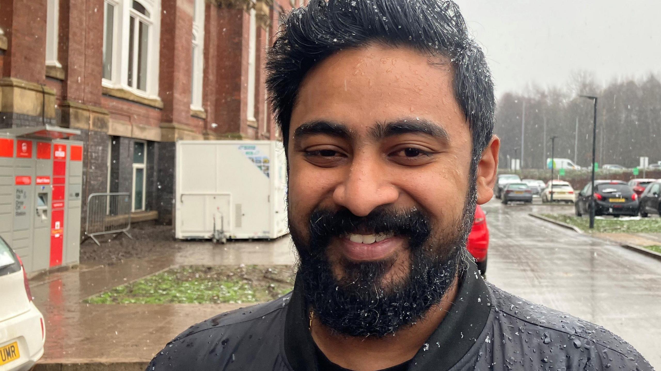 Shine Thomas stands in a black jacket outside Meadow Mill apartments as the rain falls, leaving droplets of water on his face. 