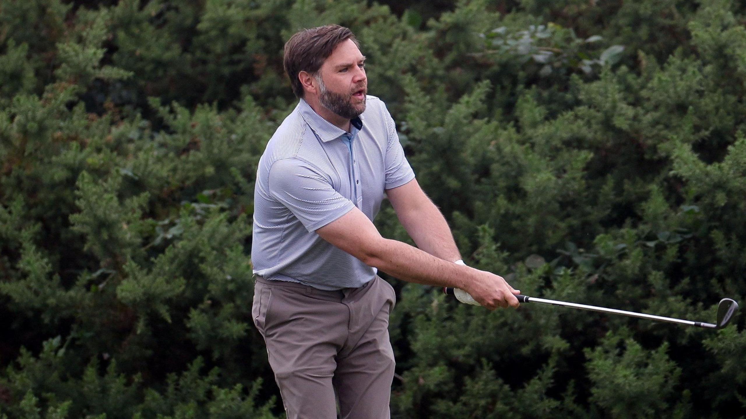 A man with brown hair and beard, wearing a light blue top an brown trousers swings a golf club with bushes behind him.