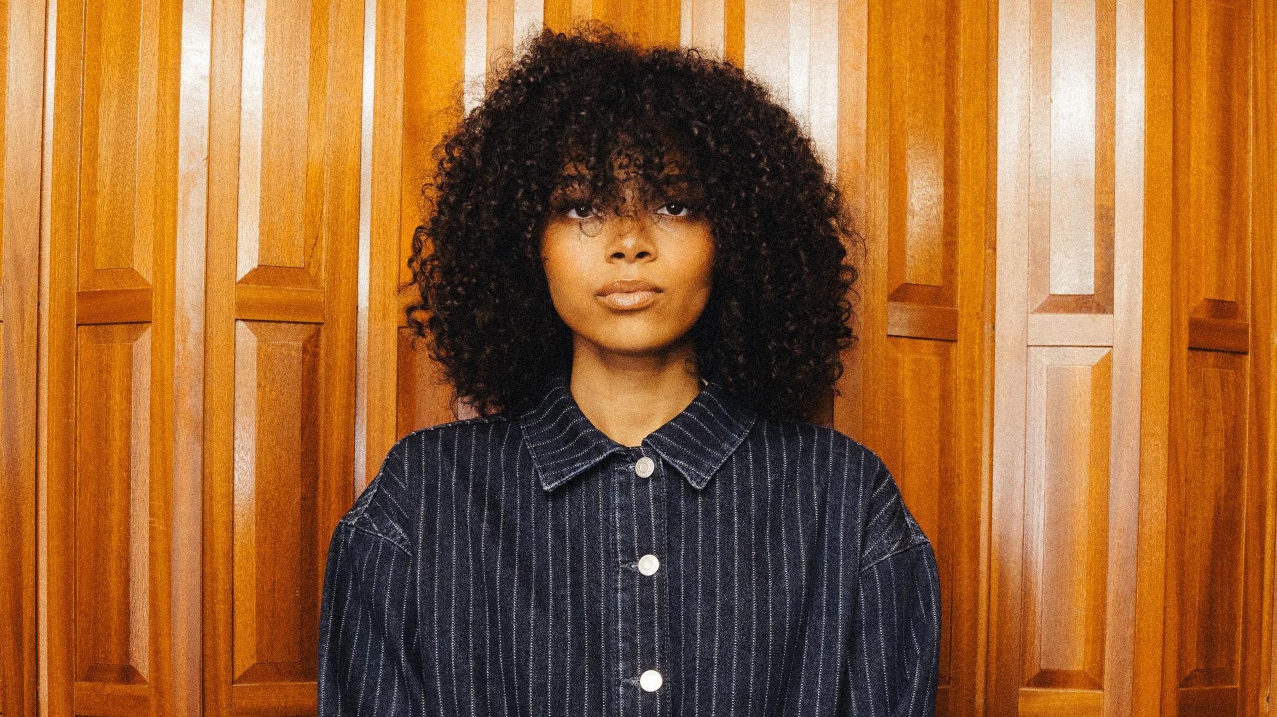 Brooke Combe is a black female artist. She has curly black hair and is wearing a dark grey pinstripe short. She standing in front of wooden panels.