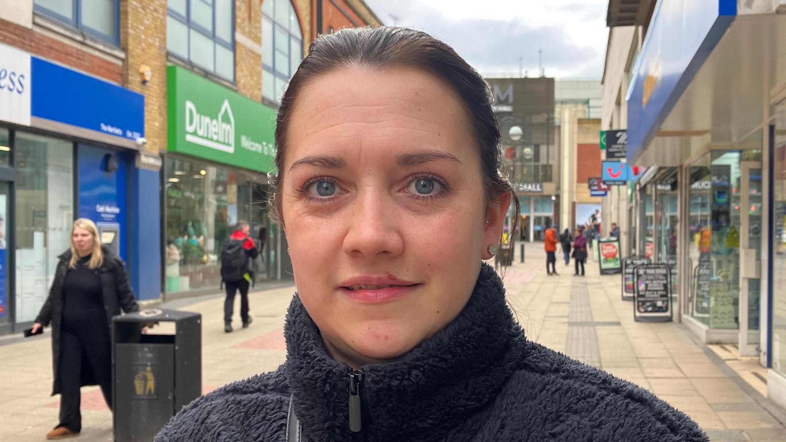 A woman with tied-up dark hair and blue eyes looking at the camera. She is wearing a dark fluffy turtle neck coat and is standing in row of shops.