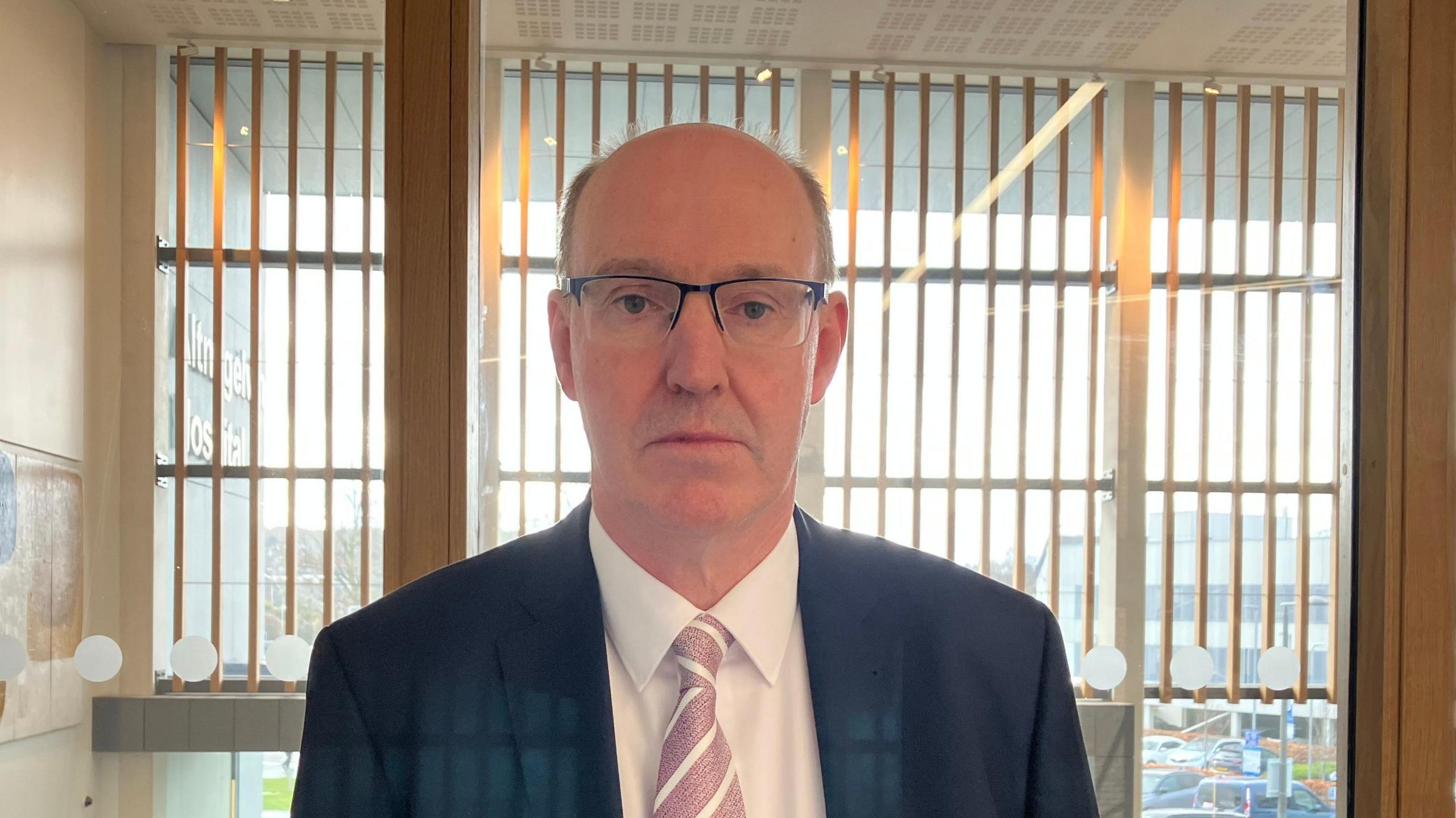 A man wearing a suit and glasses look at the camera. His tie is pink and white. Behind him are windows and wooden walls.