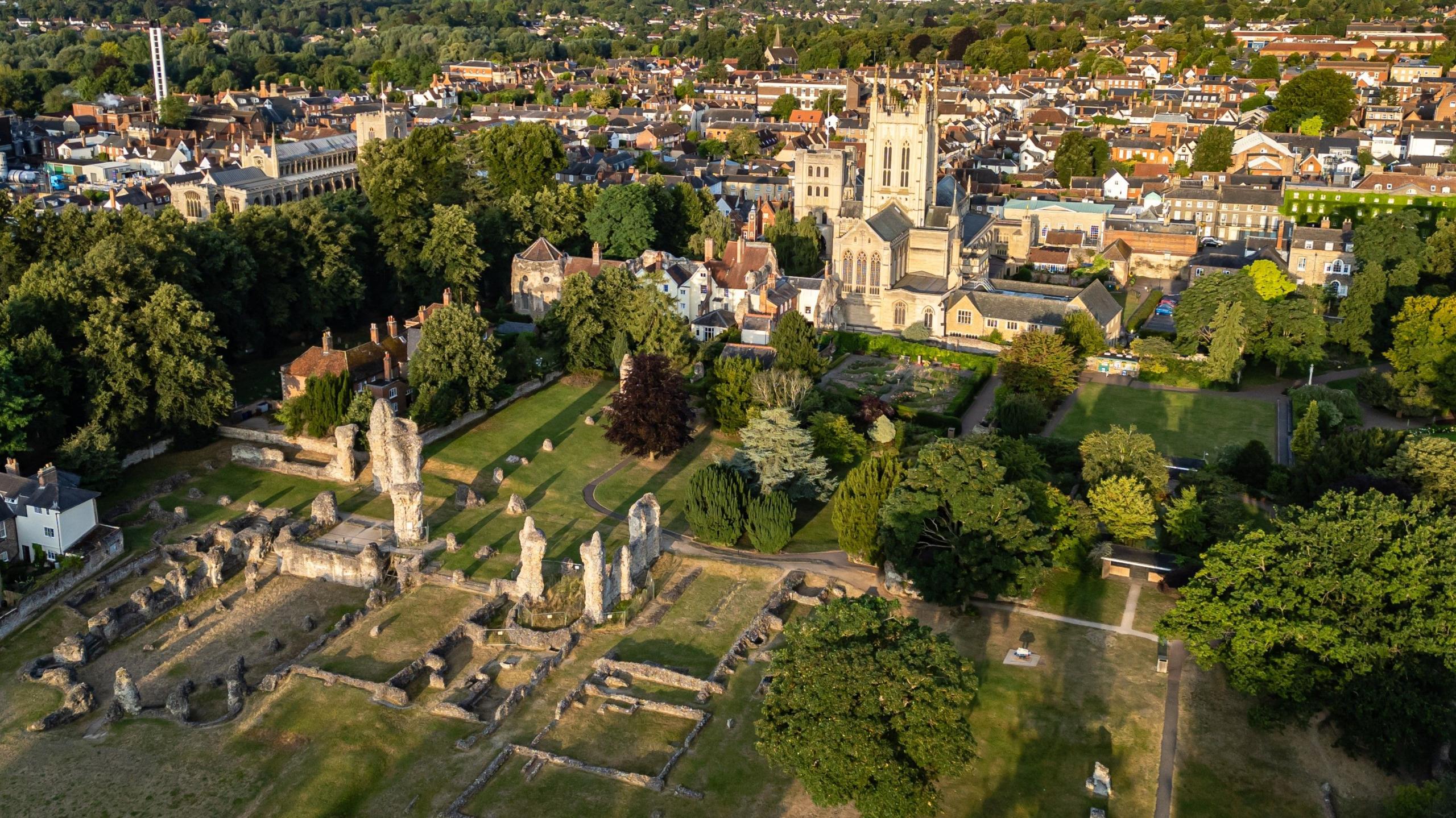 An aerial image of the sand-coloured St Edmundsbury Cathedral, which has a square tower with tall spikes at each corner. In the foreground is are grey stumps which are the ruined masonry of a medieval abbey. The site is surrounded by trees and in the background the buildings of a town centre.  