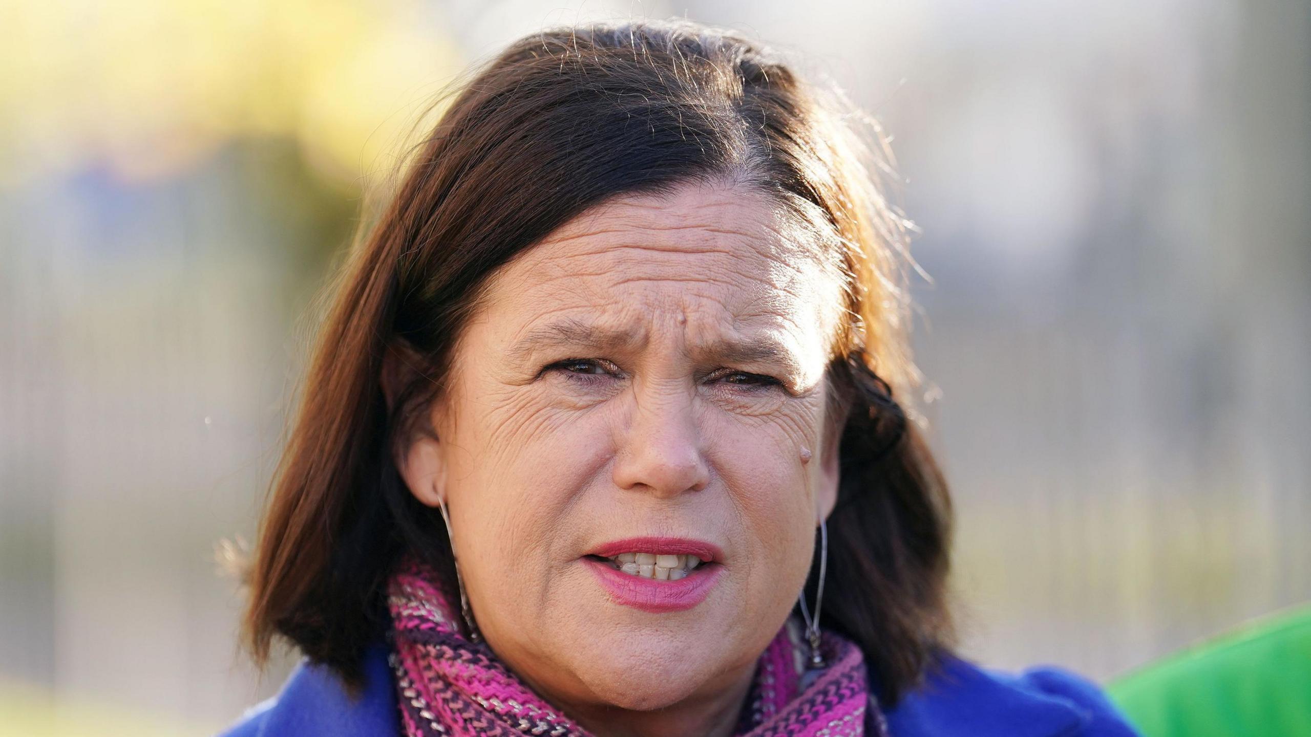 McDonald pictured with shoulder length brown hair. She stares at the camera as she wears pink lipstick, a pink scarf and a blue coat.