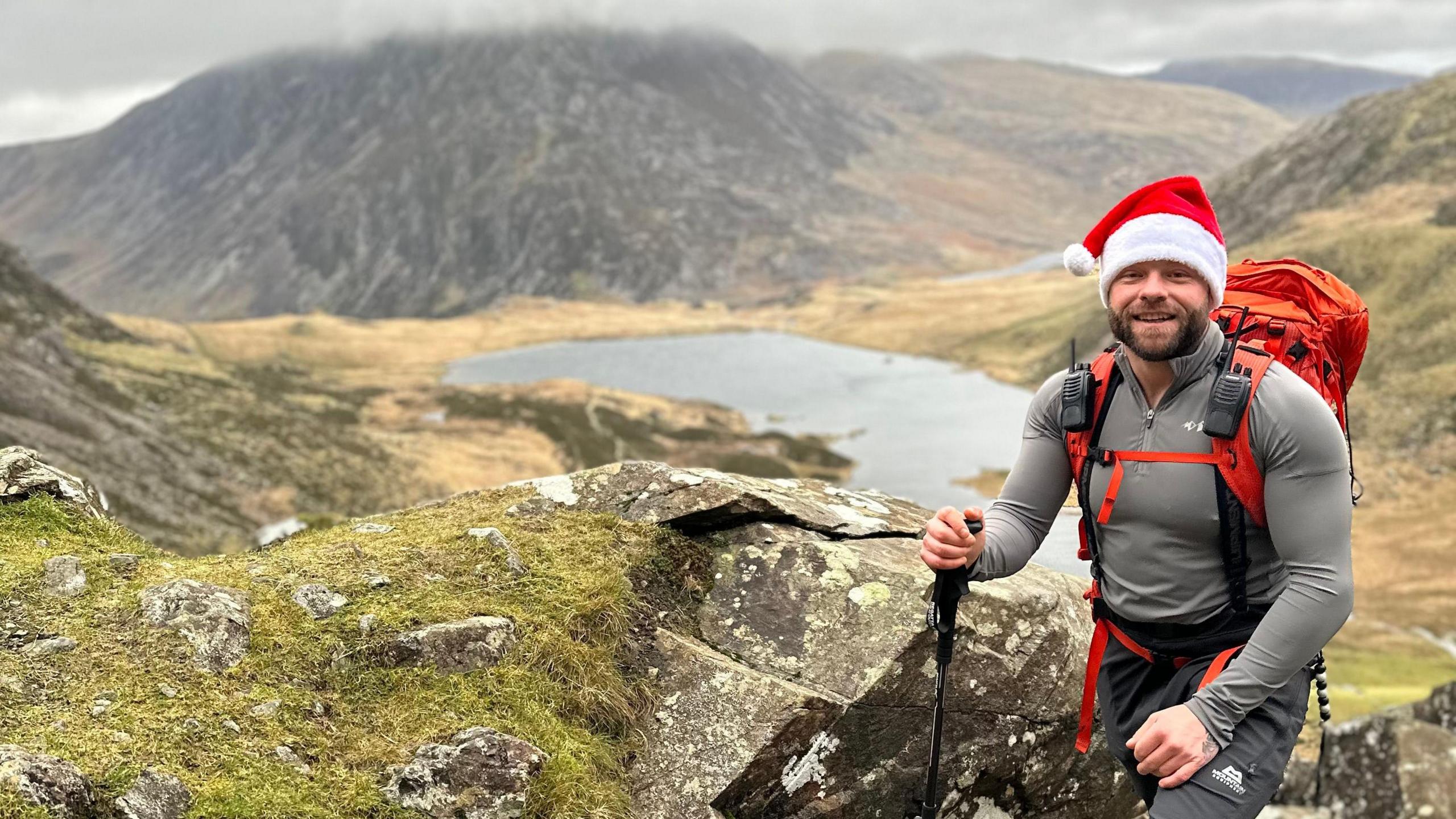 Mr Jones pictured smiling wearing a santas hat in the mountains of Eryri National Park. He carries a walking pole in his right hand and has a large backpack on his back. 