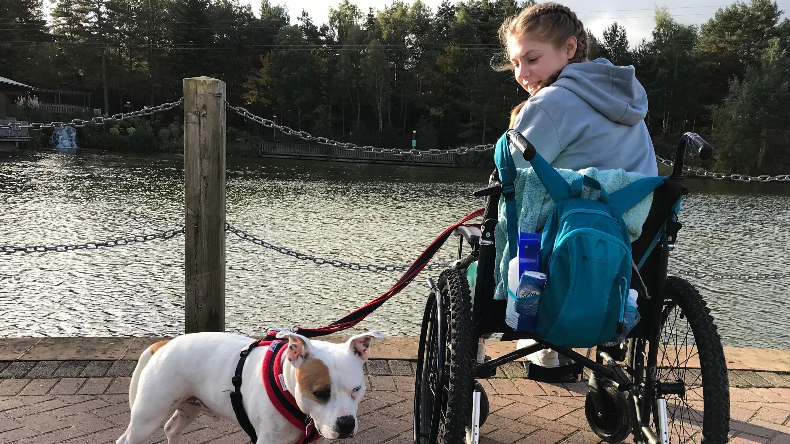 Fay Crockett is seated in a wheelchair near an iron railing overlooking a river. Her head is turned towards the camera looking at a white and brown dog she is holding with a leash