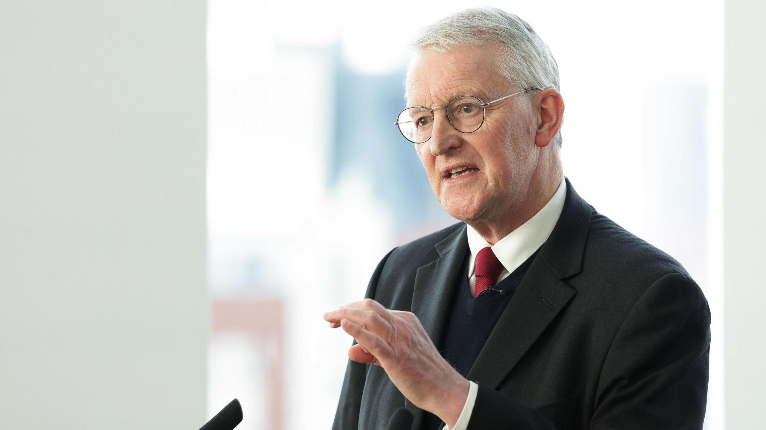 Hilary Benn speaking at a microphone. He is wearing a suit and tie with a jumper